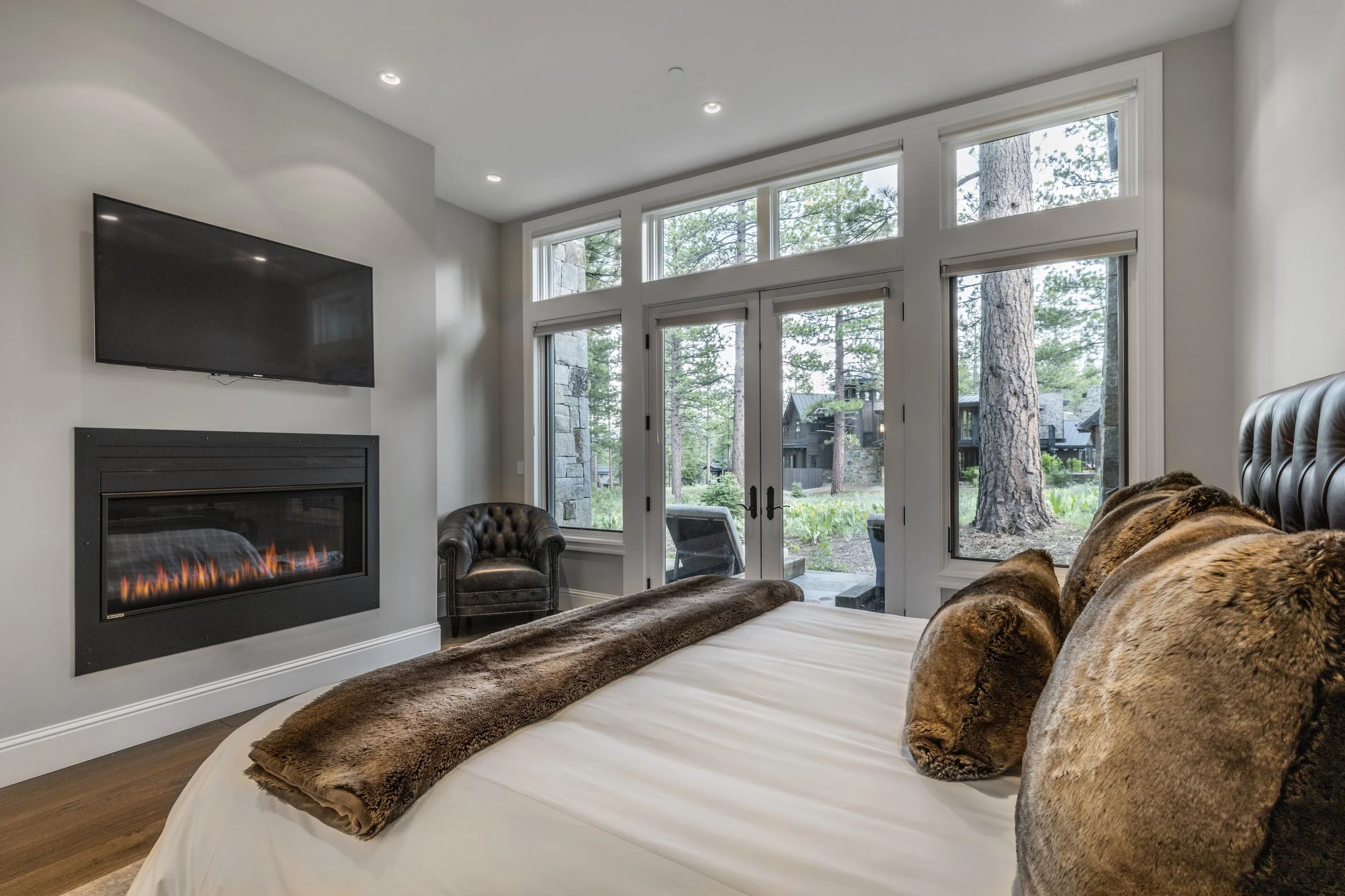 A bedroom with large windows showing a wooded outdoor view, a bed with fur pillows and throw, a wall-mounted TV above a modern fireplace, small chair, and hardwood floors.