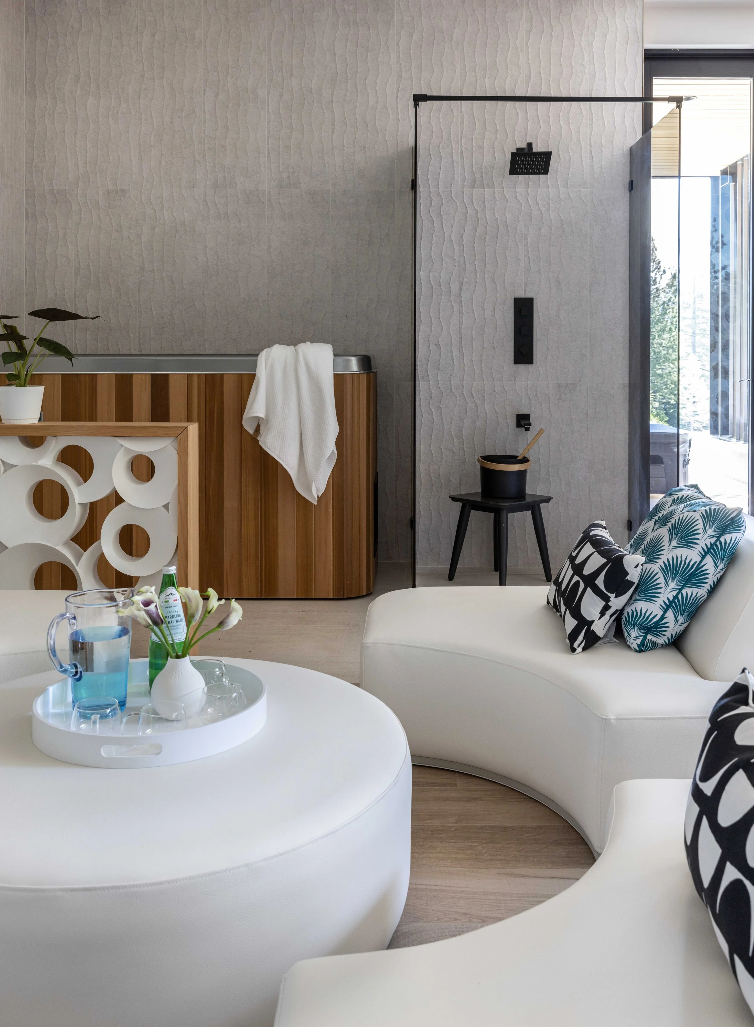 Modern living room with white curved sofas, decorative pillows, a round white coffee table with a pitcher of water and flowers, and a semi-open bathroom area with a rain shower and wooden cabinet. A large window lets in natural light.