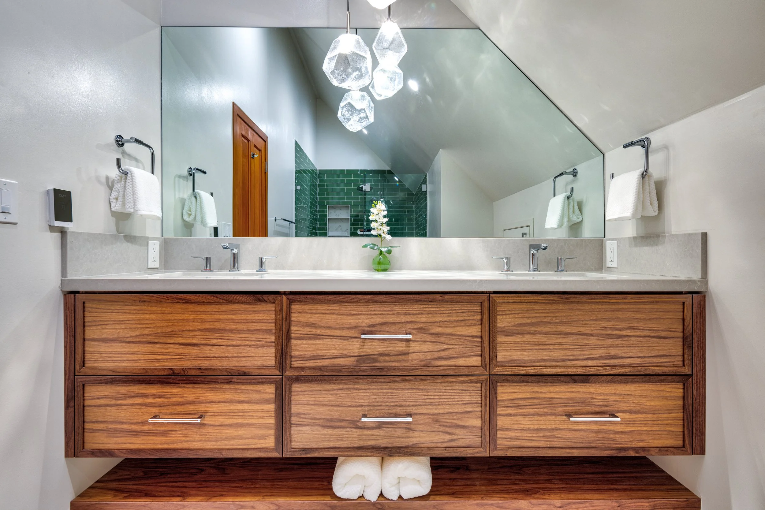Modern bathroom with double vanity, wooden drawers, large mirror, and green tiled shower area in the background.