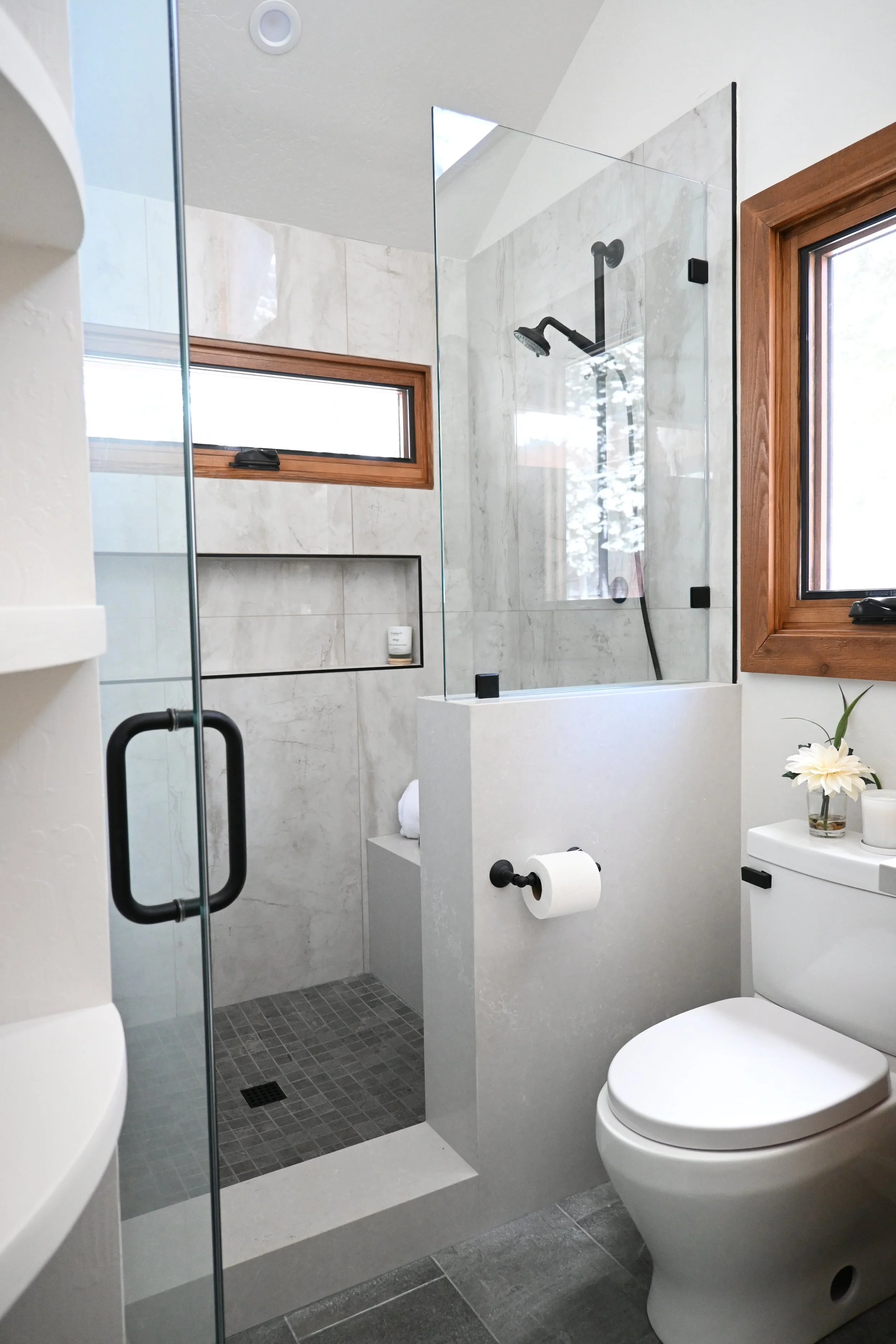Modern bathroom with a walk-in shower featuring a glass enclosure, a black shower head, and gray tiled walls. There is a toilet with a small flower arrangement on top and a window with wooden trim.