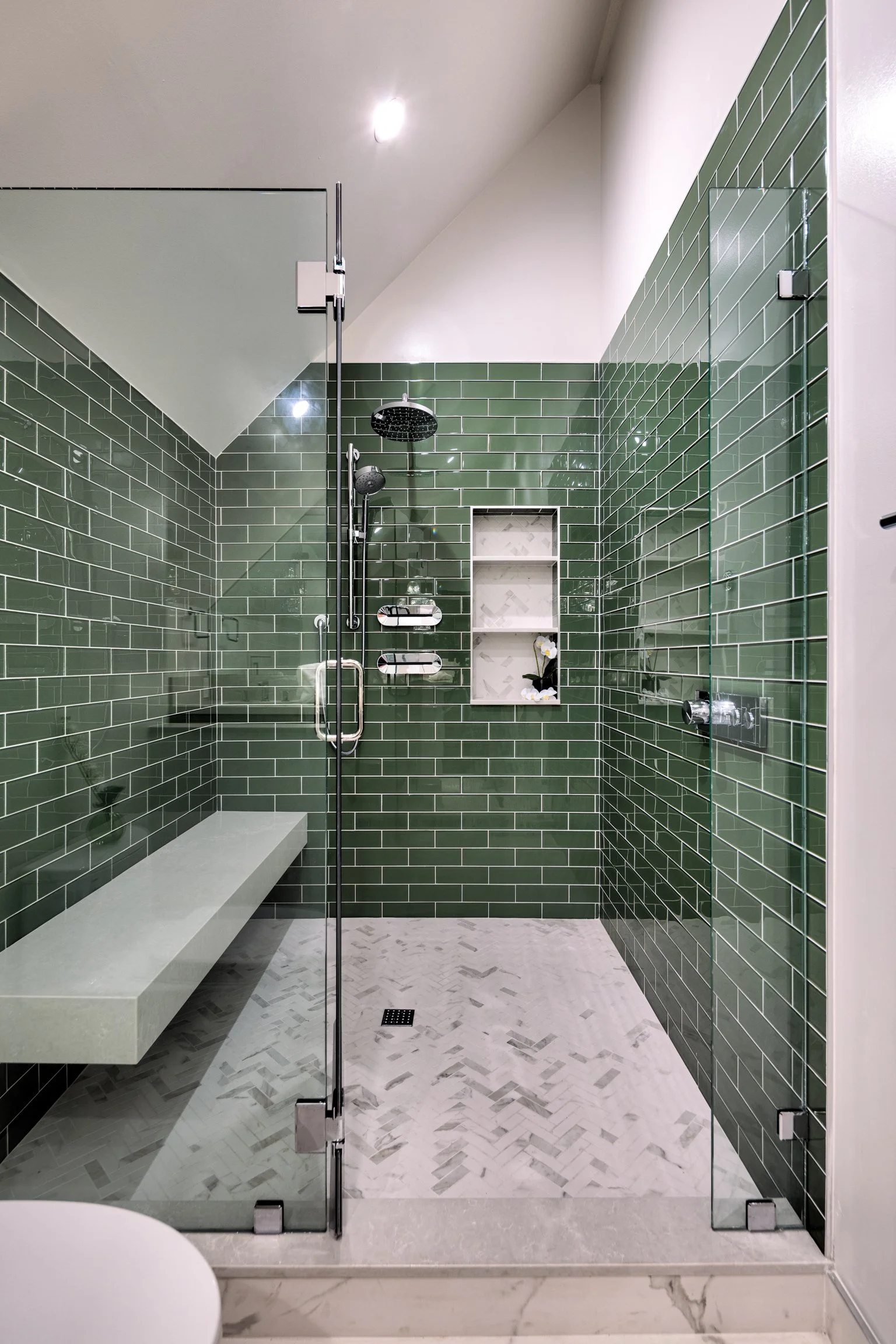 Modern shower with green subway tile walls, a two-head shower system, built-in shelves with a flower, a bench, glass enclosure, white marble floor, and recessed ceiling lighting.