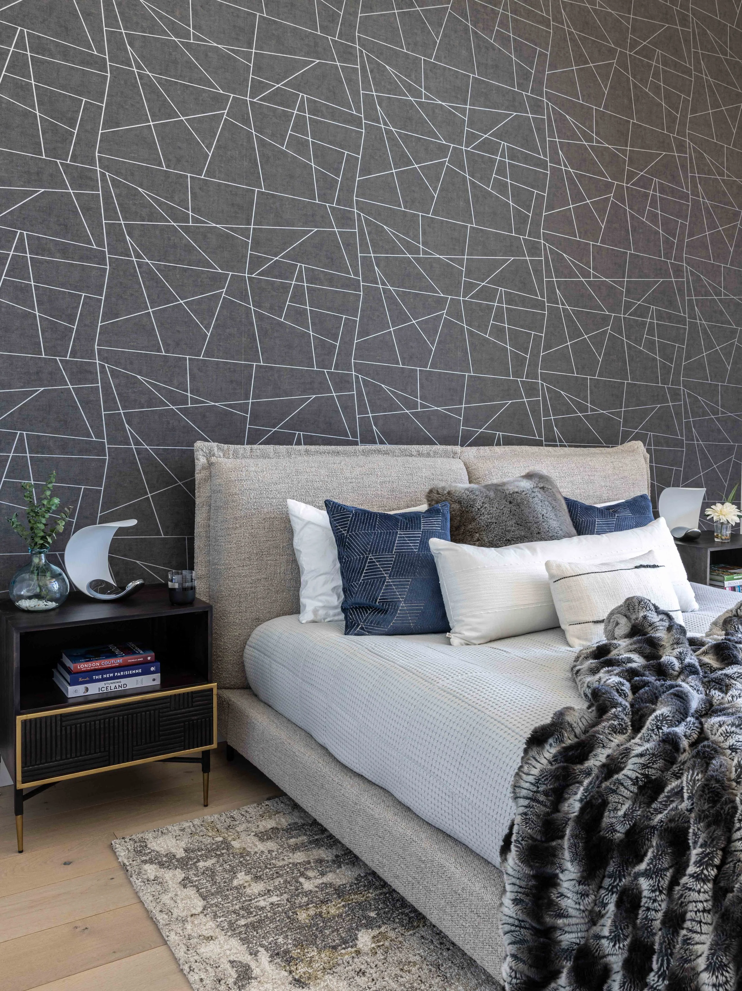 Modern bedroom with a beige upholstered bed, dark side tables, blue and white pillows, a black and white furry throw blanket, and decorative objects against a dark geometric-patterned wall.