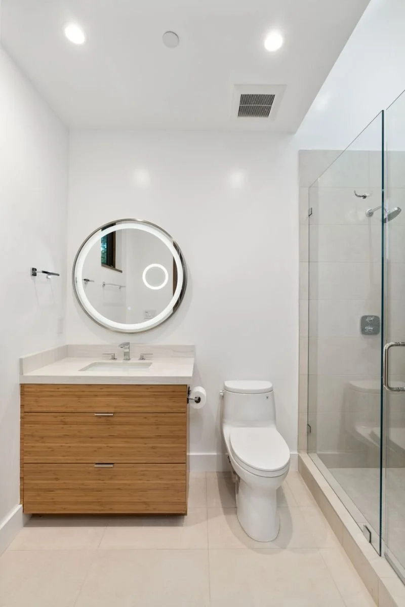 Modern bathroom with white walls, a round mirror, a wooden vanity with a white countertop, a toilet, and a glass shower enclosure.