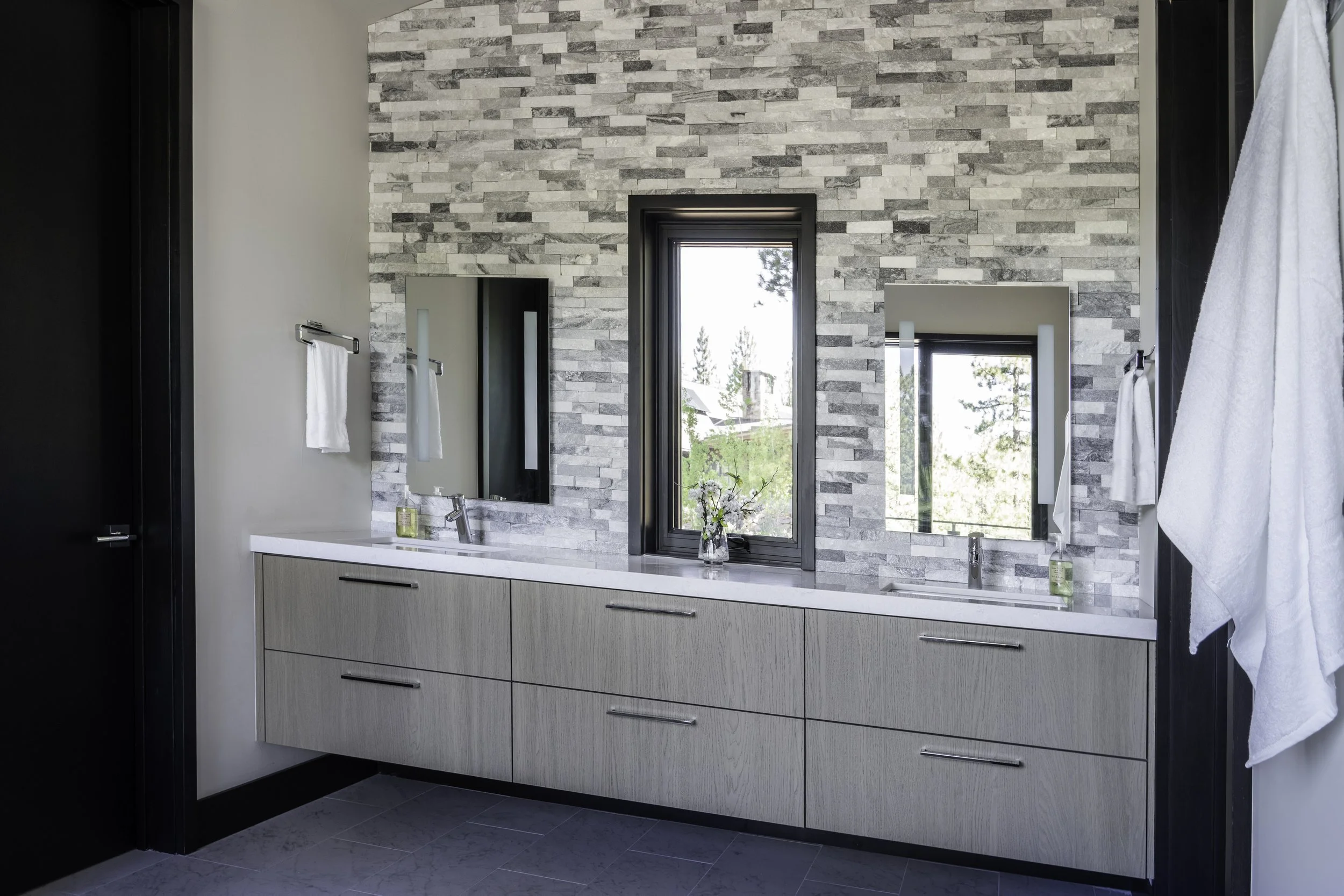 Modern bathroom with a double vanity, light wood cabinets, white countertop, two mirrors, stone accent wall, and two small windows with outdoor views.