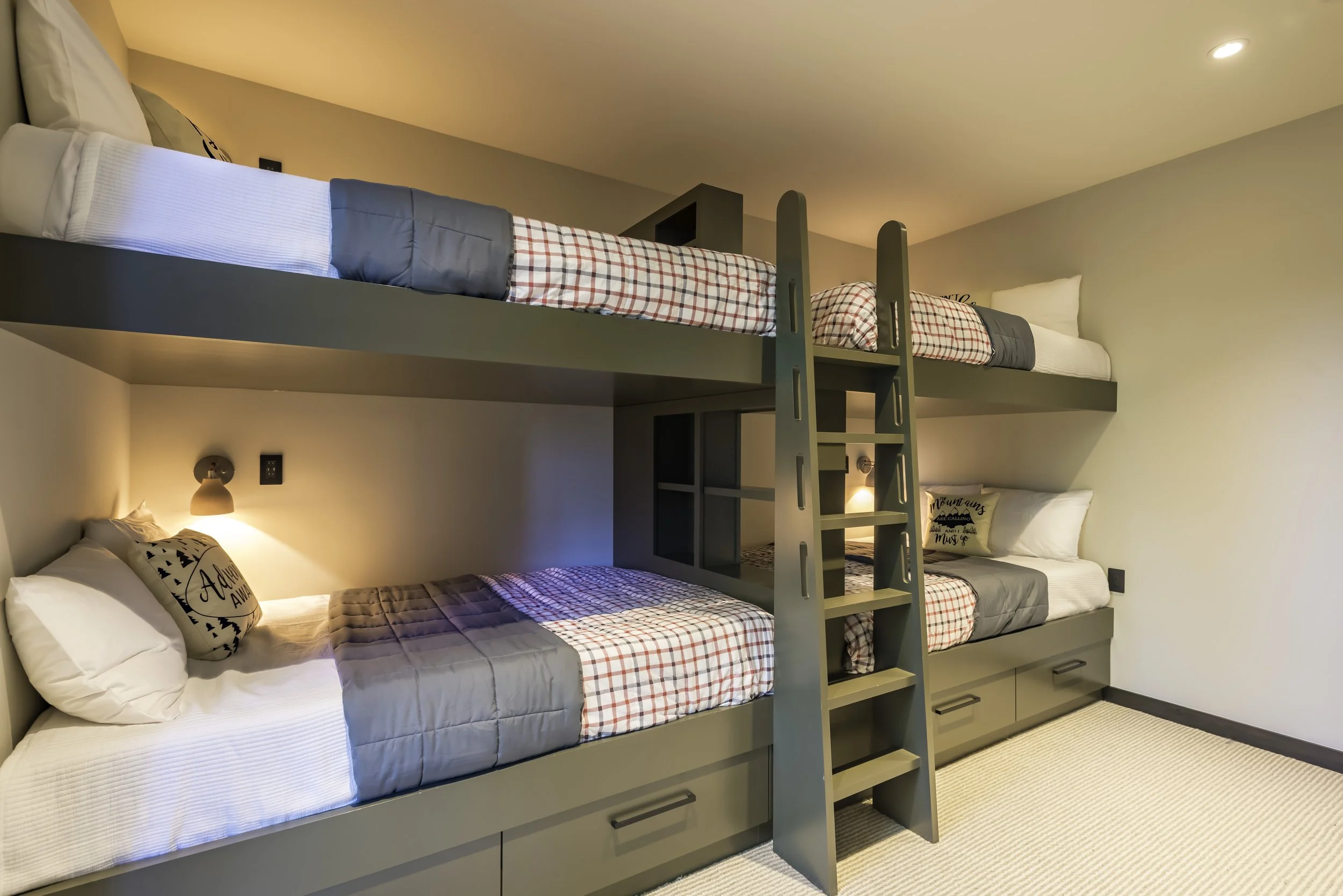 A room with two sets of bunk beds, each with a lower double bed and an upper single bed, with gray bedding, plaid blankets, and decorative pillows. The beds have built-in drawers underneath and a small ladder for access to the top bunks.