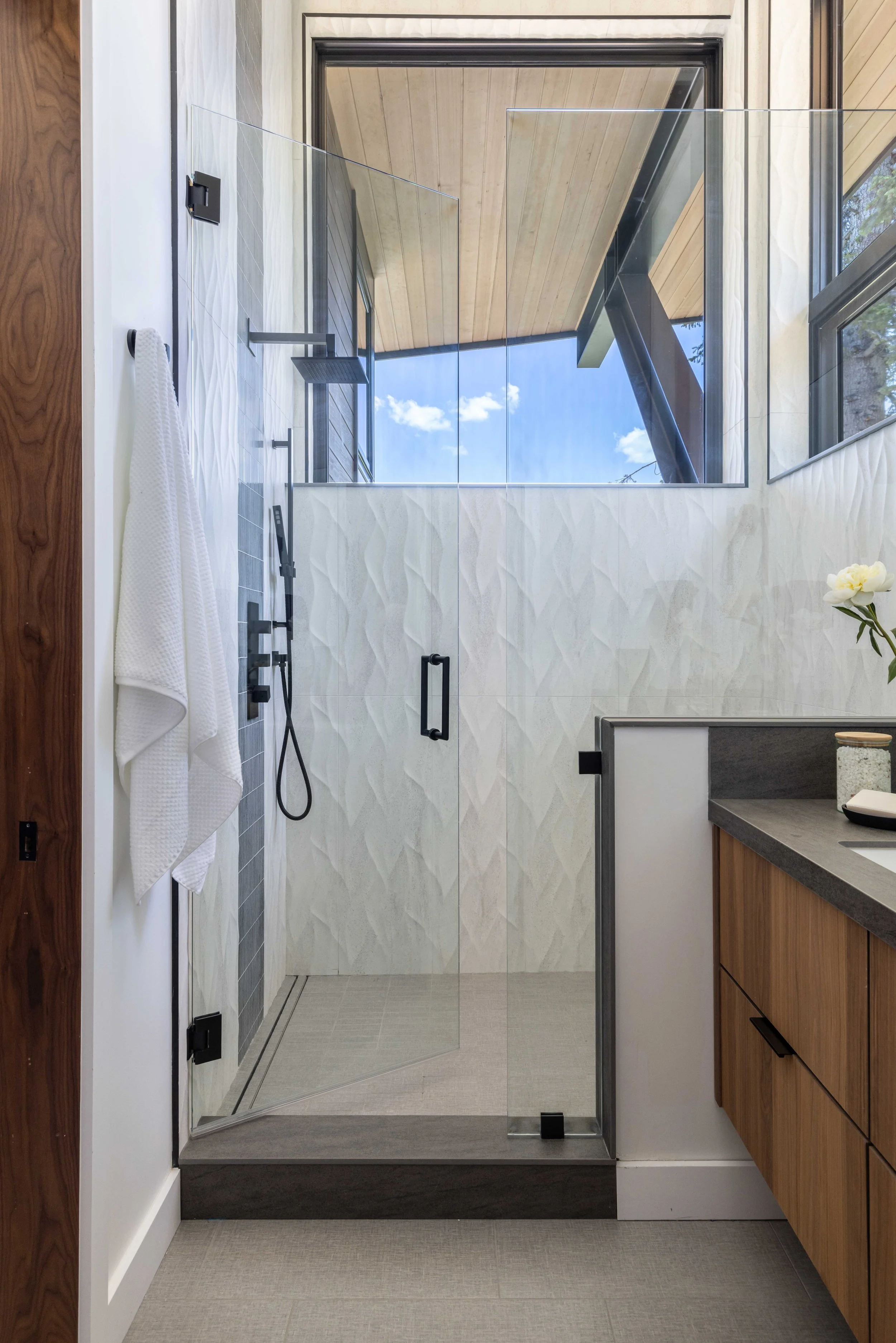Modern bathroom with a glass shower enclosure, wooden cabinets, and a large window showing a blue sky with clouds.