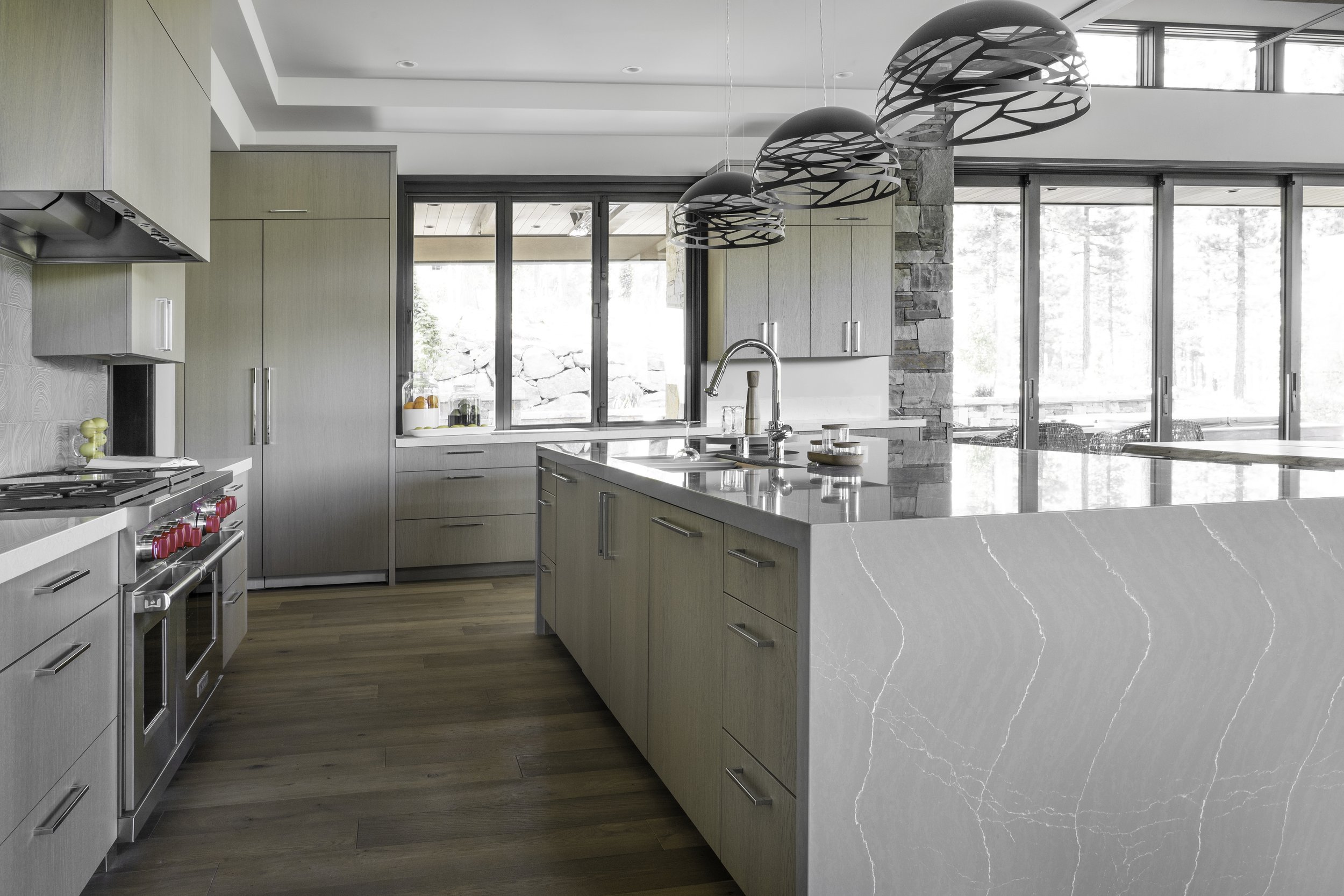 Modern kitchen with light wood cabinets, stainless steel appliances, and an island with a light-colored marble countertop. Large windows and sliding glass doors bring in natural light, overlooking a stone yard outside. Three decorative pendant lights