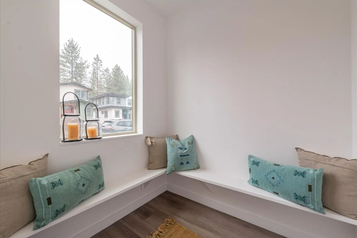 A corner window seat with pillows, a large window showing houses and trees outside, and candles on the window ledge.