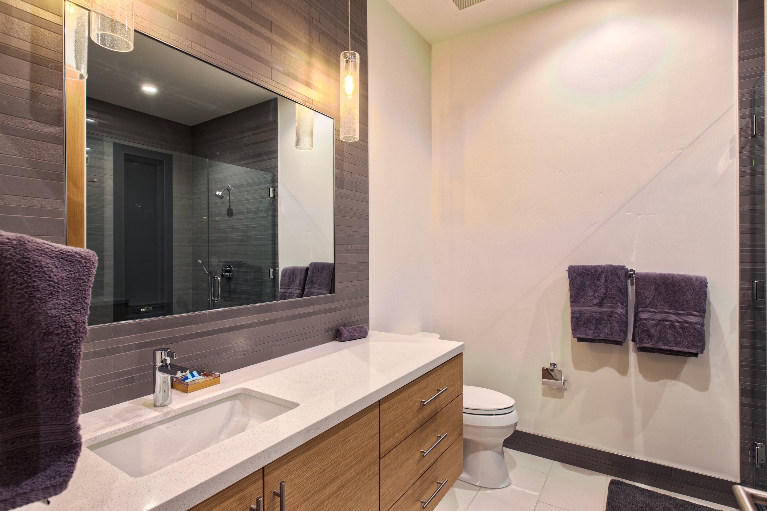Modern bathroom with a large mirror, a white sink on a wooden cabinet, a toilet, and two purple towels hanging on the wall. The shower area with glass doors is visible in the reflection.