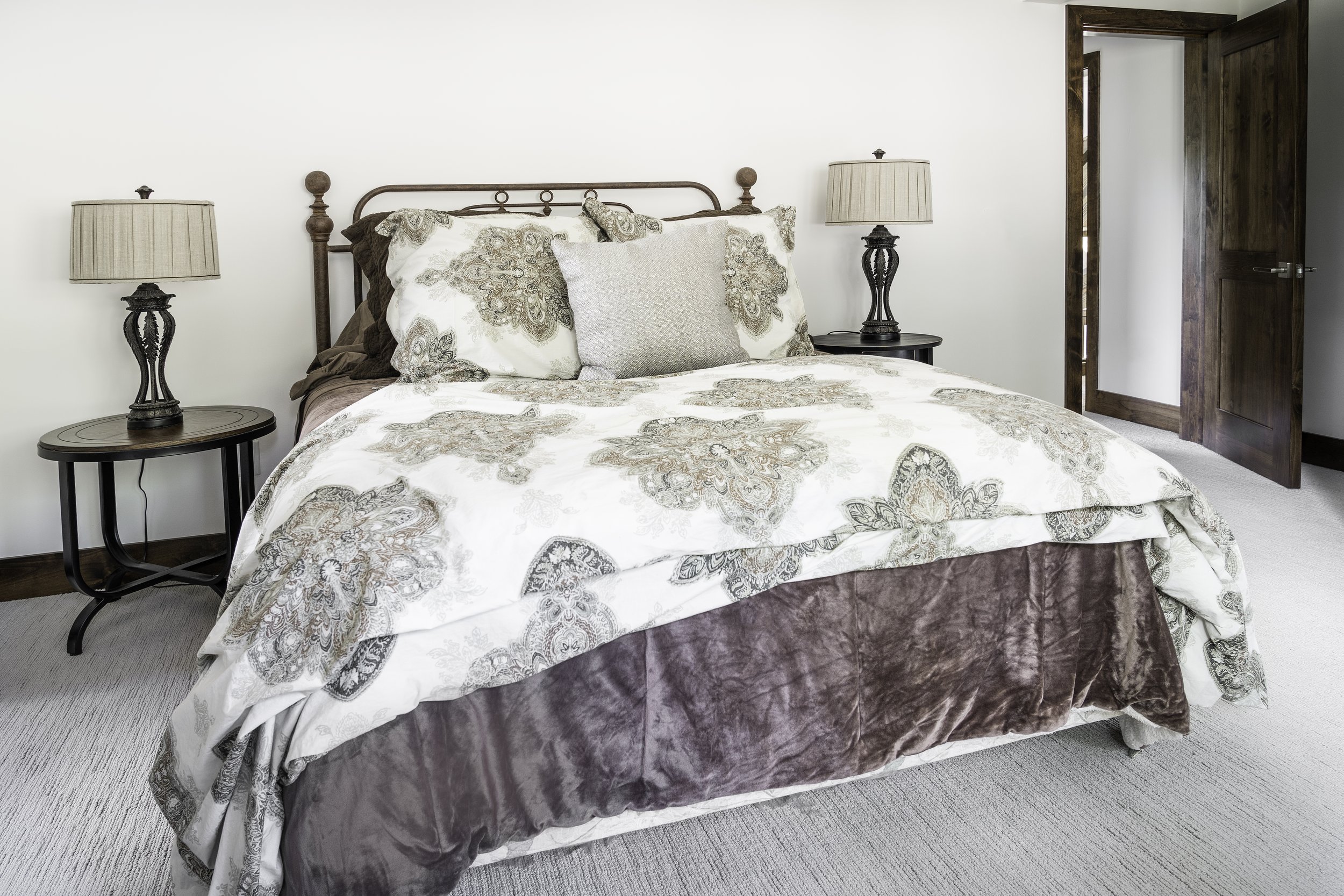 A neatly made bed with floral-patterned bedding and multiple pillows, flanked by two nightstands with matching lamps, in a bedroom with wooden doors and trim.