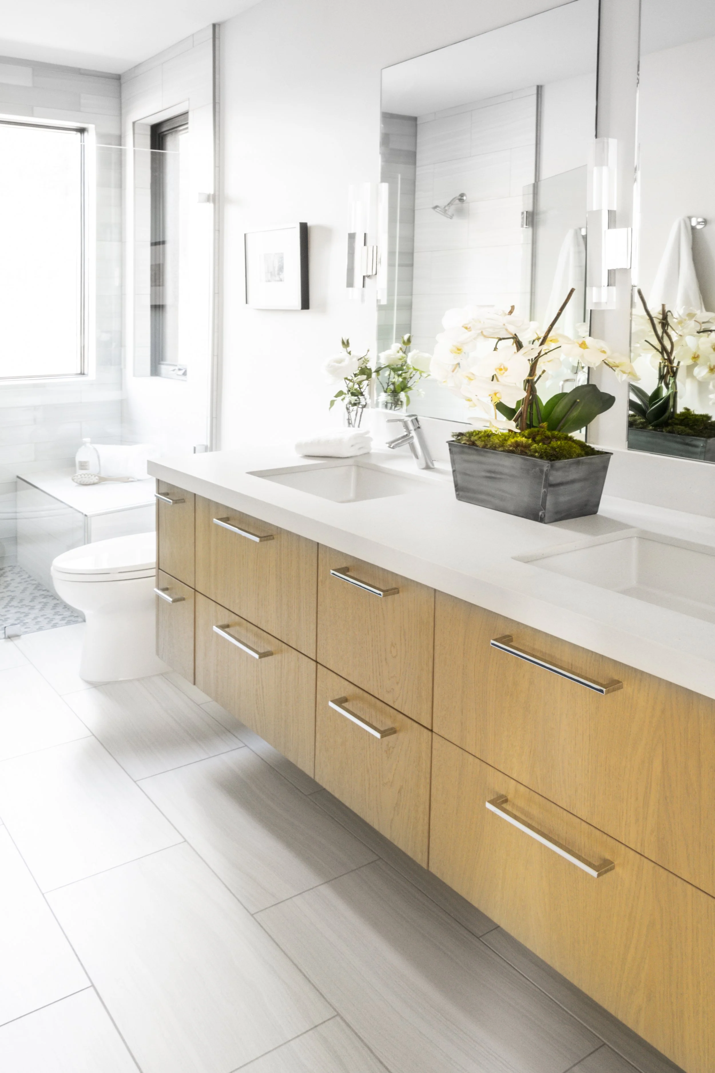 Modern bathroom with double vanity, white countertop, wooden cabinets, large mirror, orchid plant, walk-in shower, and toilet with a window