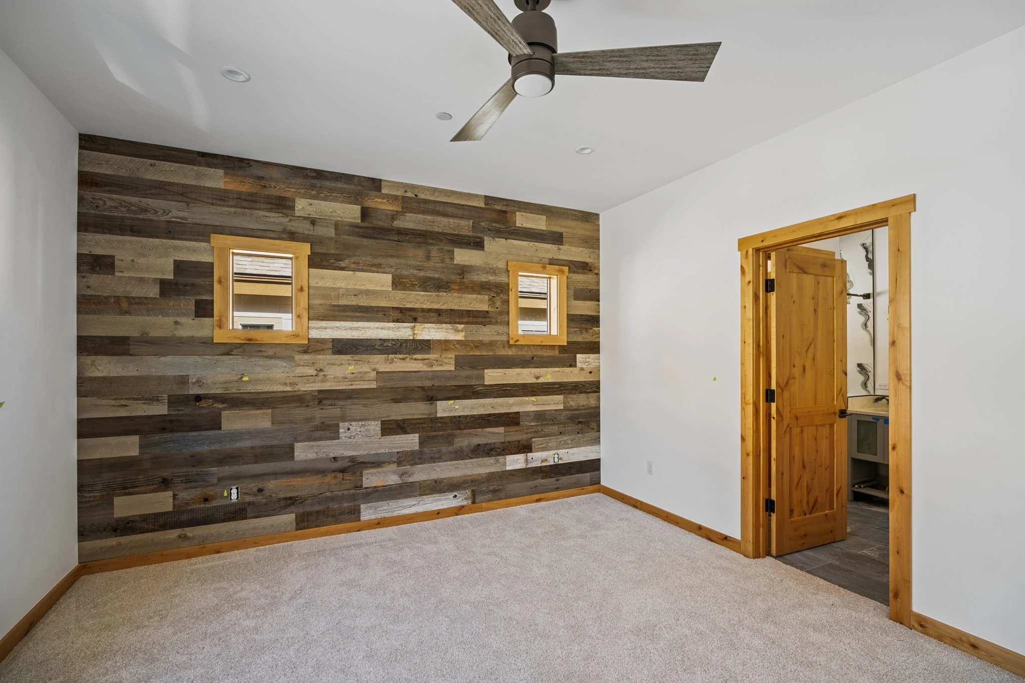 Empty room with a wooden accent wall, two small windows, a ceiling fan, and a doorway leading to another room with wooden trim.
