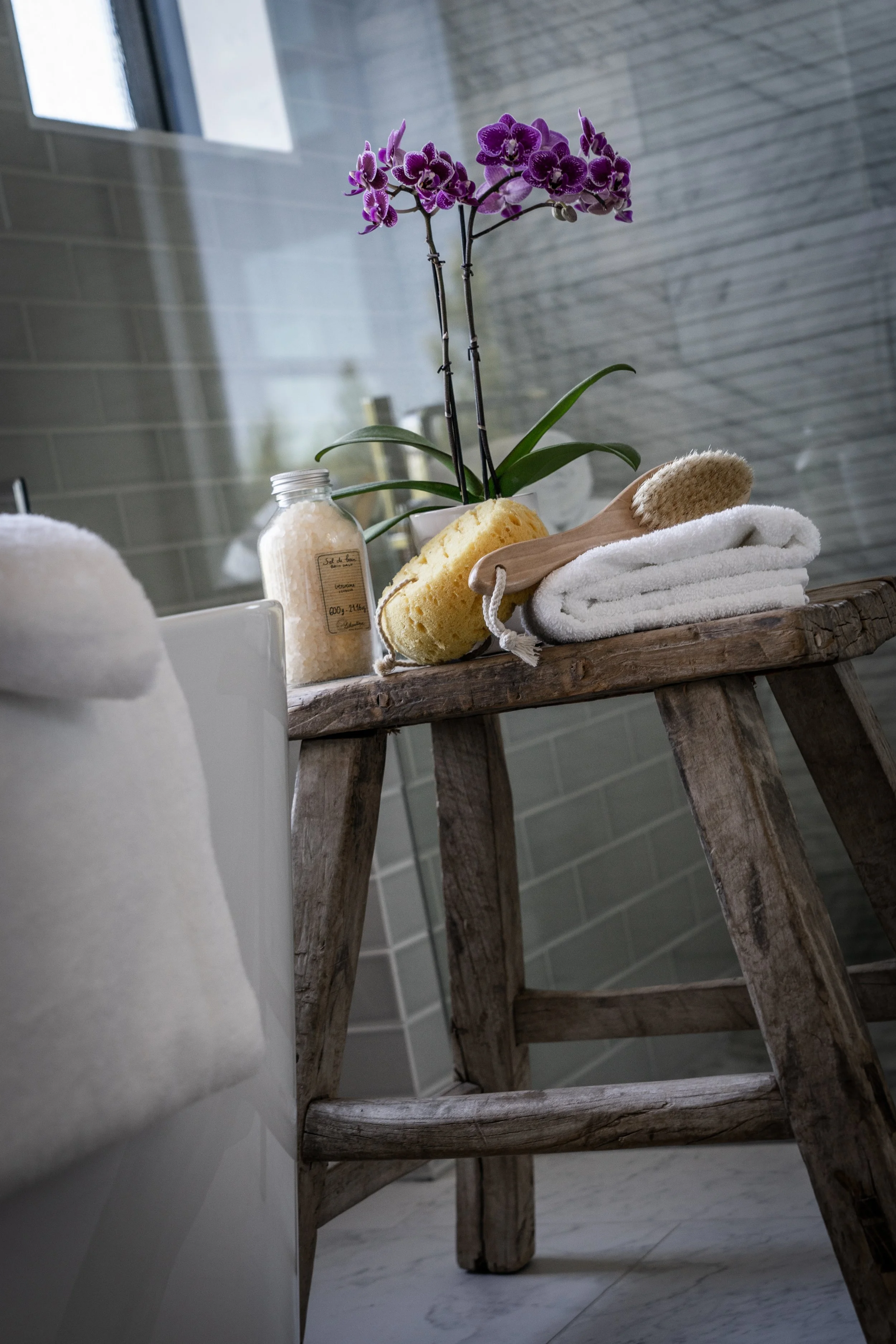 A wooden stool in a bathroom holds a potted purple orchid, a sponge, a white towel, a bath brush, and a jar of bath salts.