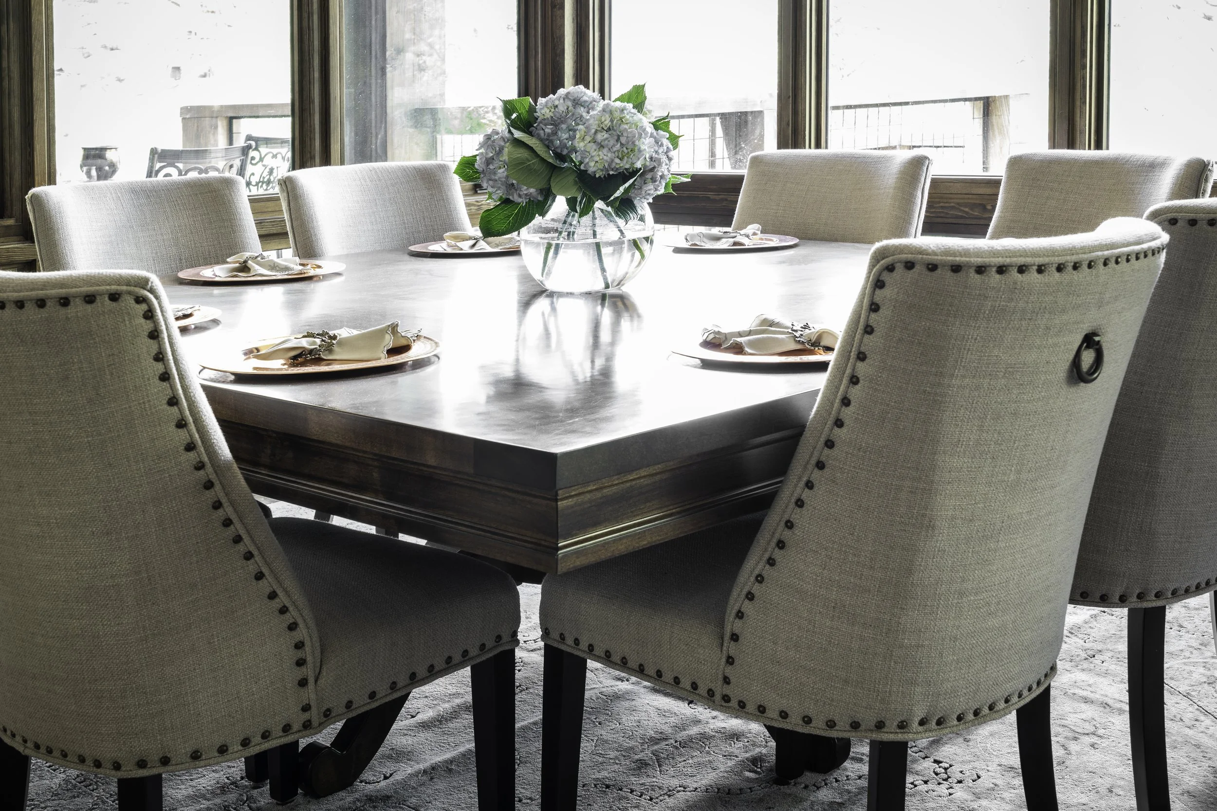 Round wooden dining table with six beige upholstered chairs surrounding it in a well-lit room with large windows. A glass vase with white hydrangea flowers is in the center of the table, with place settings including plates, napkins, and silverware.