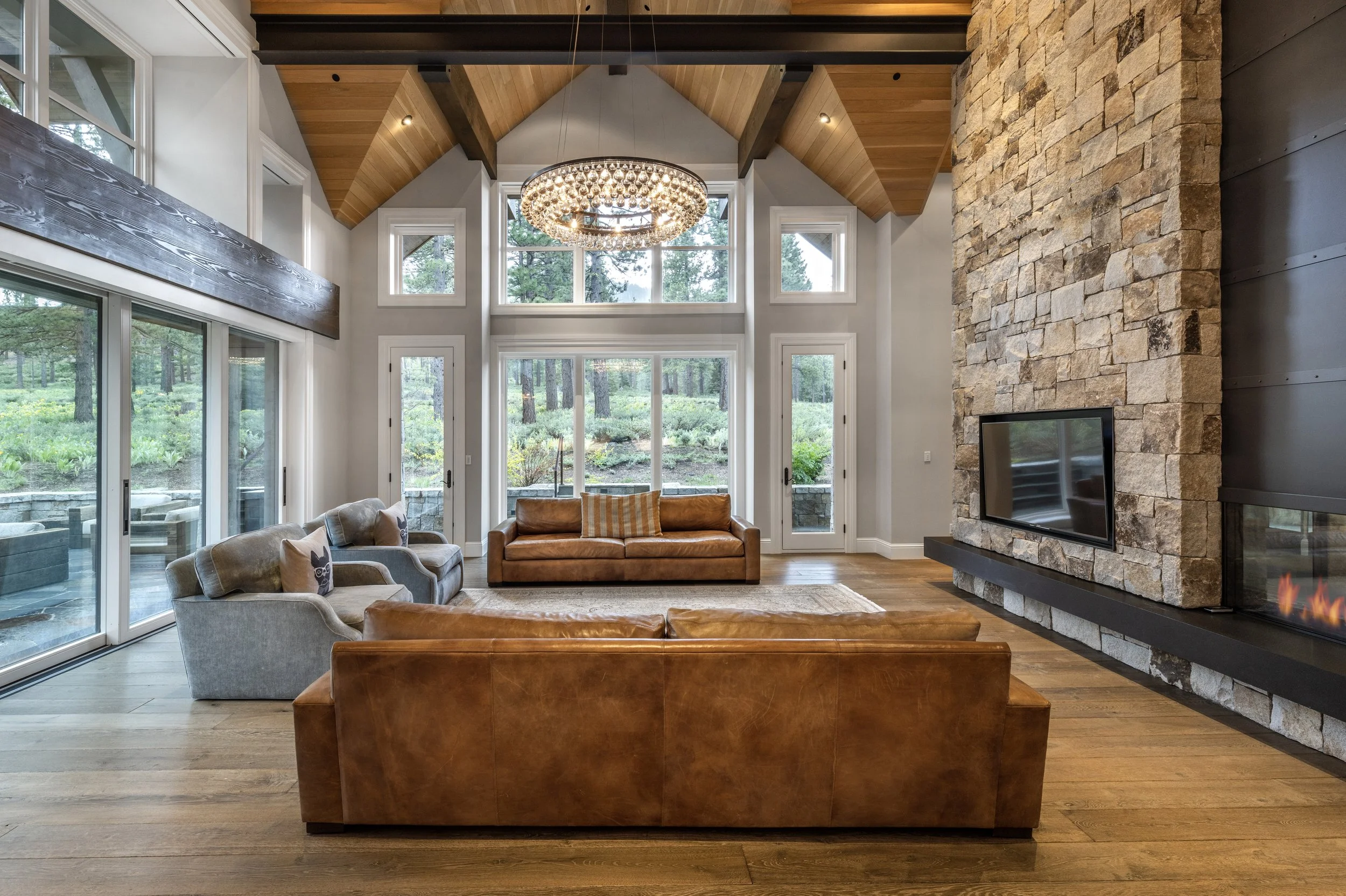 Spacious living room with large windows, high vaulted ceiling with wood beams, a stone fireplace, leather and fabric sofas, a rug, a chandelier, and a view of greenery outside.