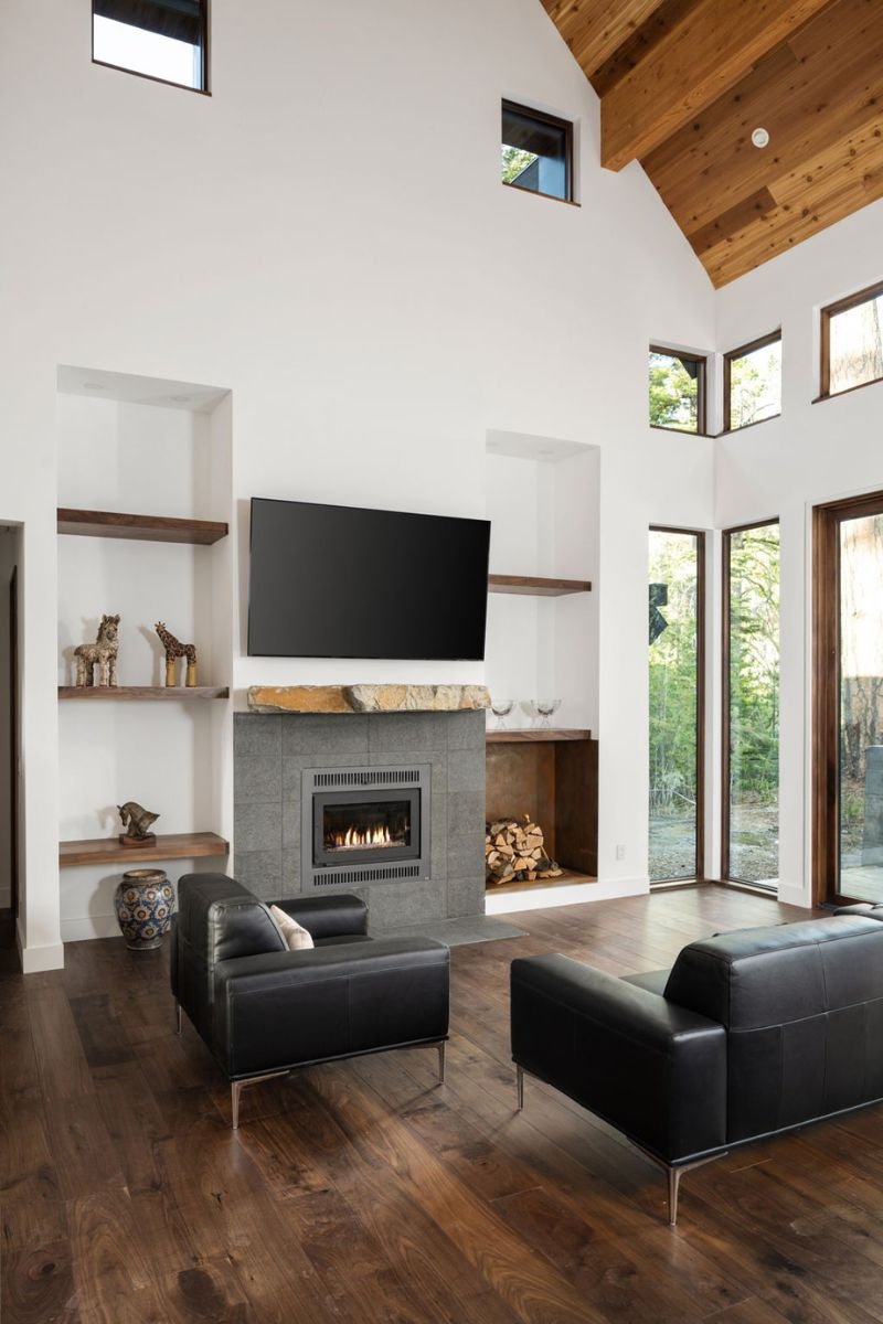 Modern living room with a ceiling of wood planks, white walls, large windows, a wall-mounted TV, a fireplace with a stone mantel, and black leather sofas.