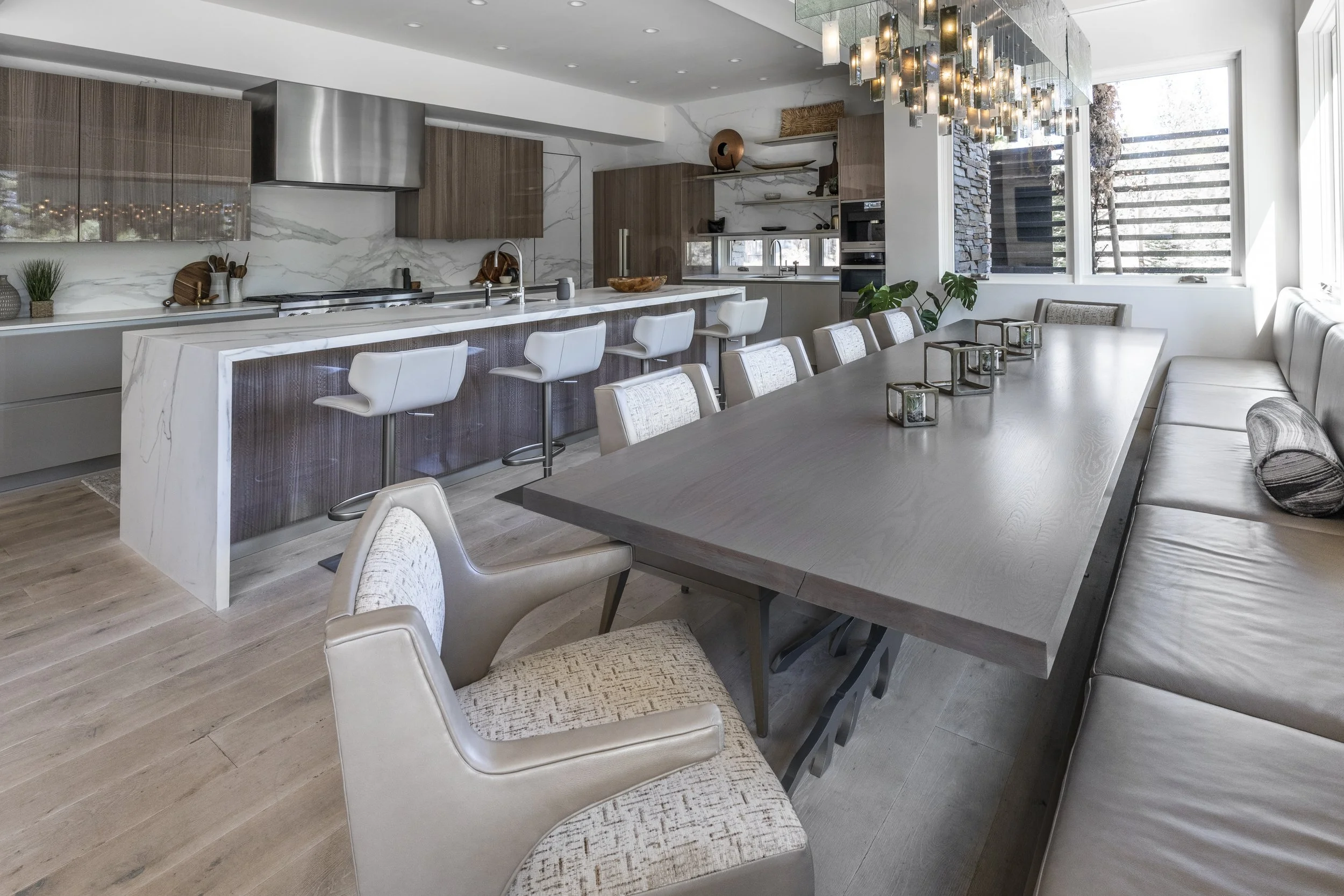 Modern kitchen and dining area with white marble countertops, wooden cabinets, and a long dining table with beige chairs. Large window with outdoor view and decorative lighting fixture hanging from the ceiling.