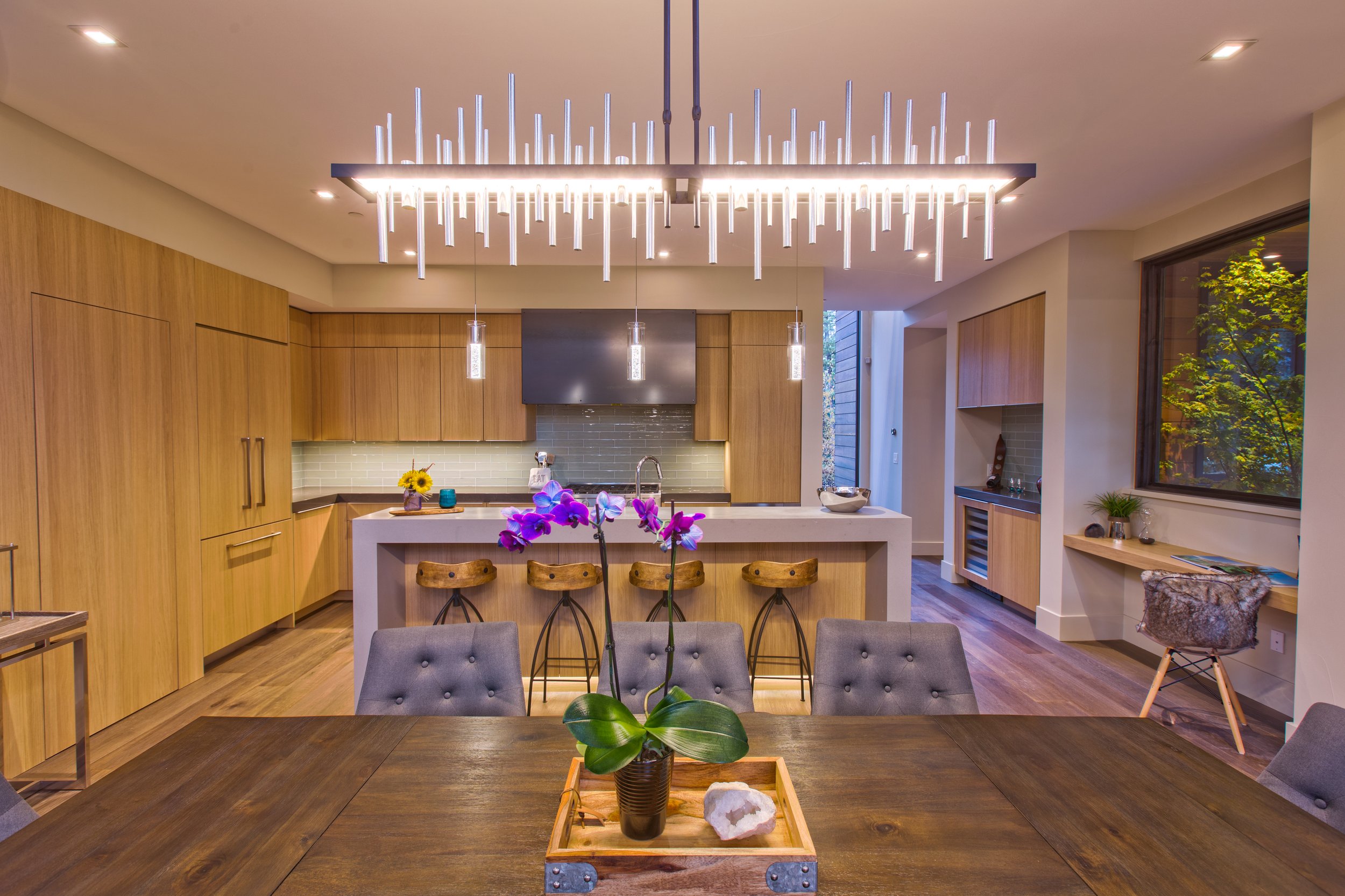 Modern kitchen with a wooden dining table, gray chairs, a white kitchen island with purple orchids, hanging pendant lights, wooden cabinets, and large window with trees outside.