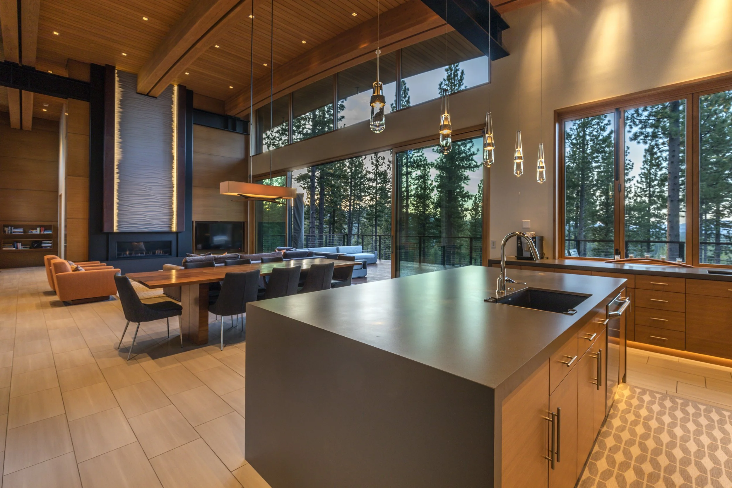 Modern open-concept kitchen and living area with large windows overlooking trees, featuring a kitchen island with a sink, a dining table with chairs, a sitting area with a fireplace, and contemporary lighting fixtures.