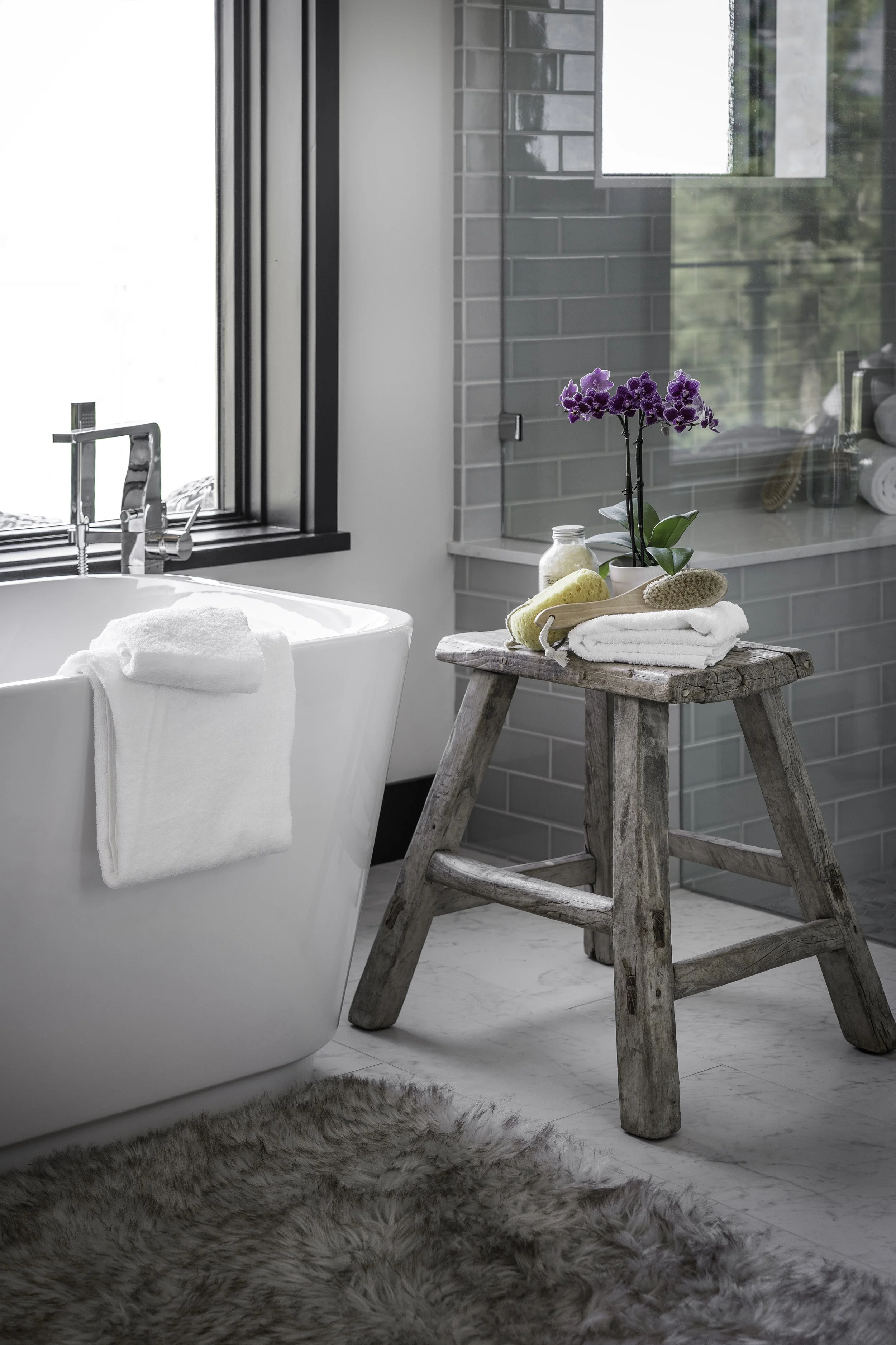 Bathroom with a freestanding bathtub, a wooden step stool holding a potted orchid, towels, brushes, and a window with outside view.