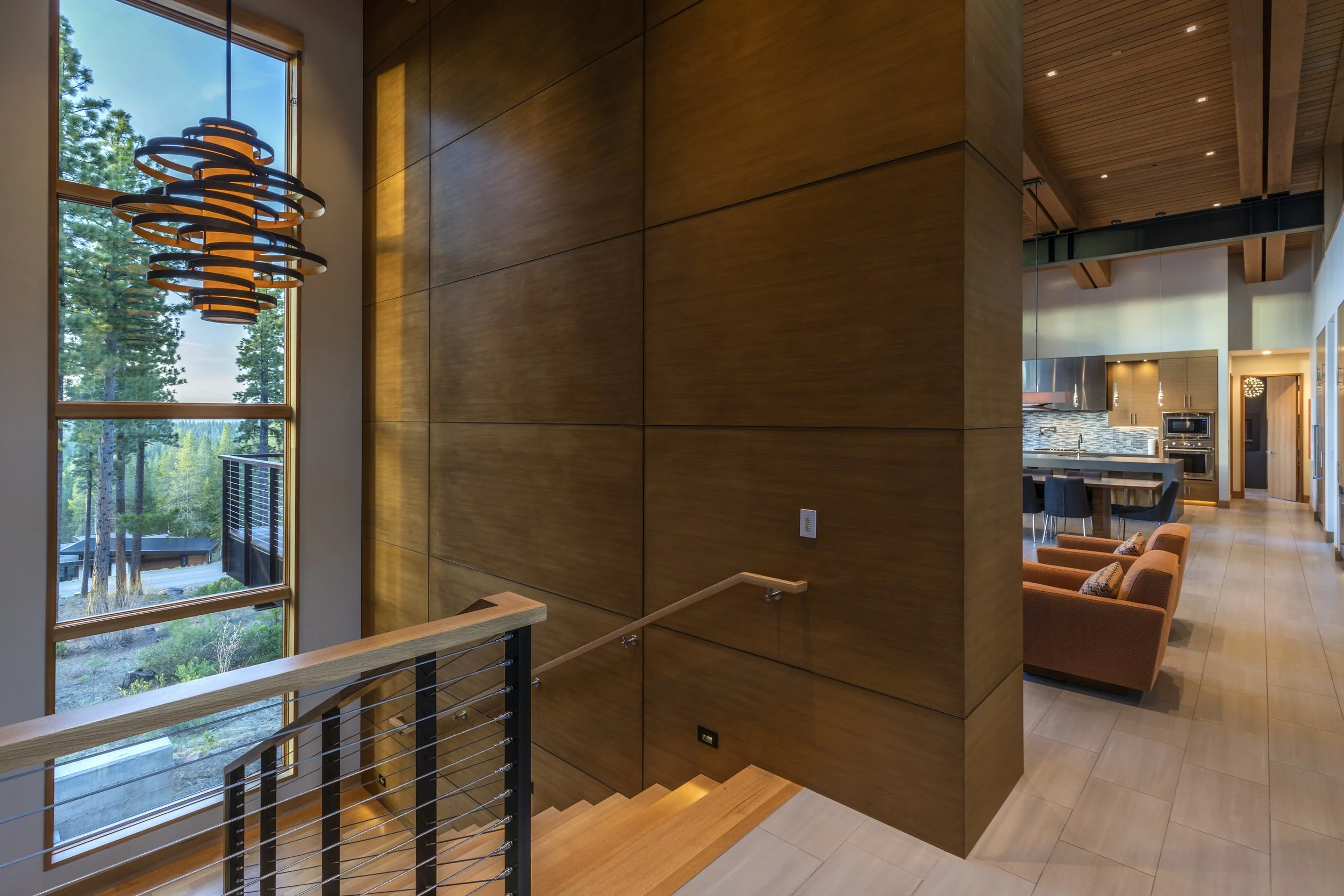 Interior view of a modern home with a wooden staircase, large windows showing trees outside, a hanging light fixture, and a kitchen area with an island and seating.