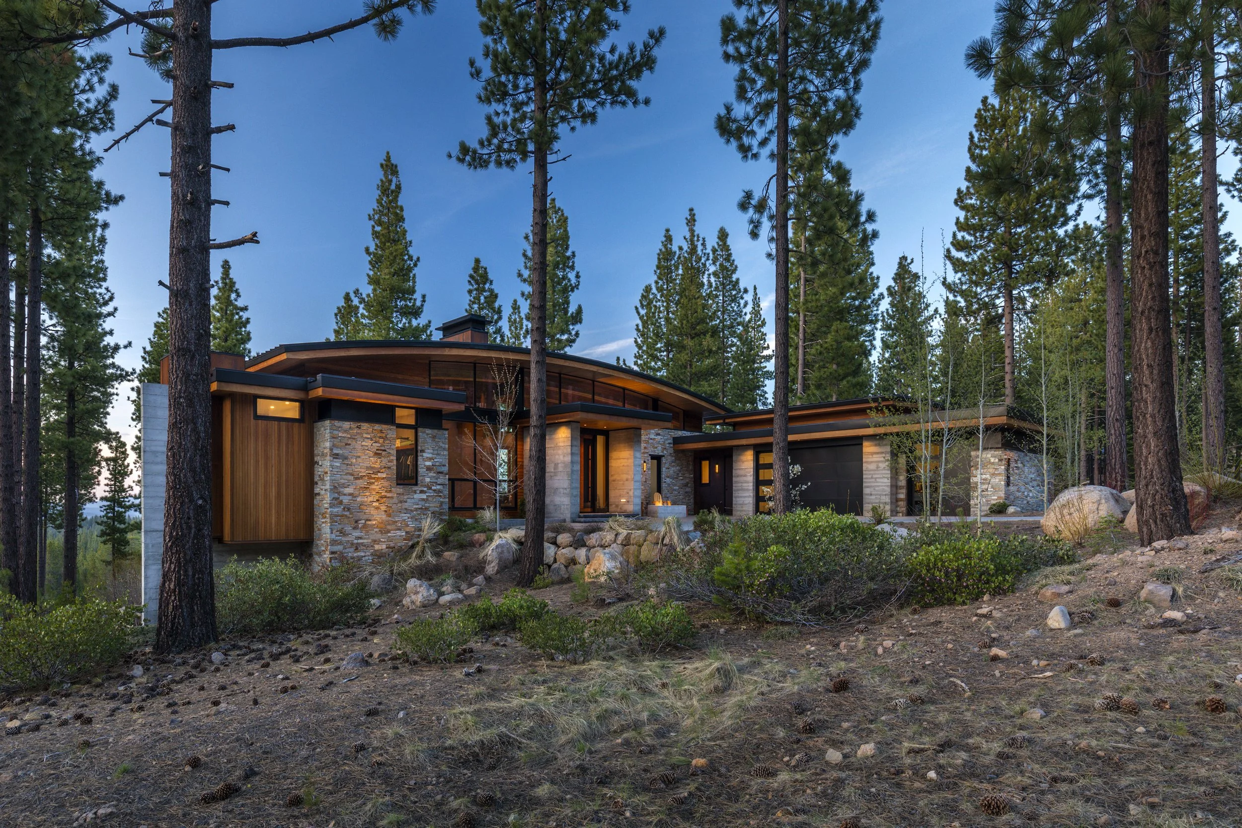 Modern house with stone and wood exterior surrounded by pine trees and nature at dusk.