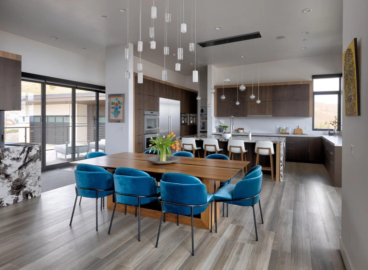Modern open-concept kitchen and dining area with a wooden table surrounded by blue velvet chairs and a kitchen with dark wood cabinets, white marble countertops, and pendant lighting.