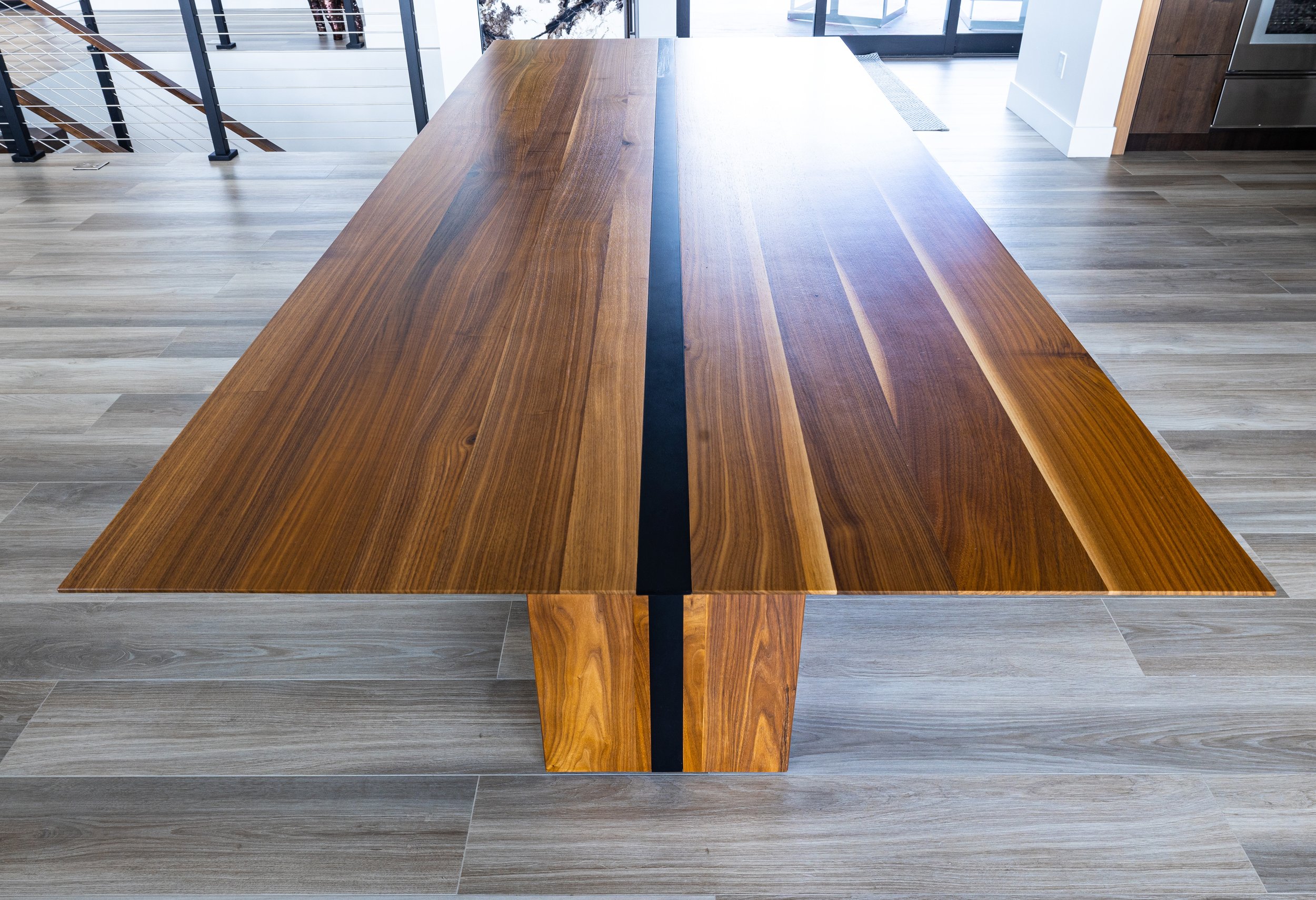 A wooden dining table with a black strip in the center, situated on a light-colored wood floor in a modern kitchen or dining area.