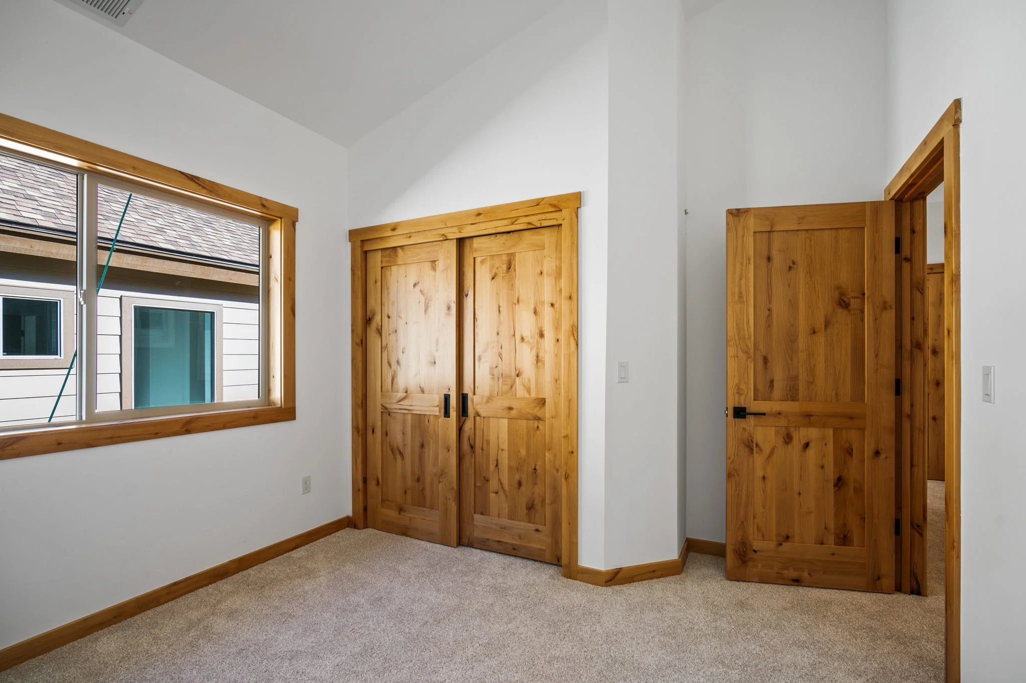Empty room with white walls, carpeted floor, wooden trim, a window on the left, a double wooden closet door, and an open wooden door leading to another room.