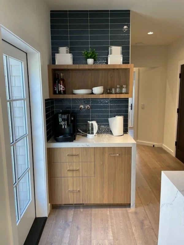 Kitchen kitchenette with wooden cabinets, a coffee machine, electric kettle, and paper towel holder on a white marble countertop. Open wooden shelf with white dishes, glasses, and bottles. Blue tiled backsplash, white potted plant, and decorative sto
