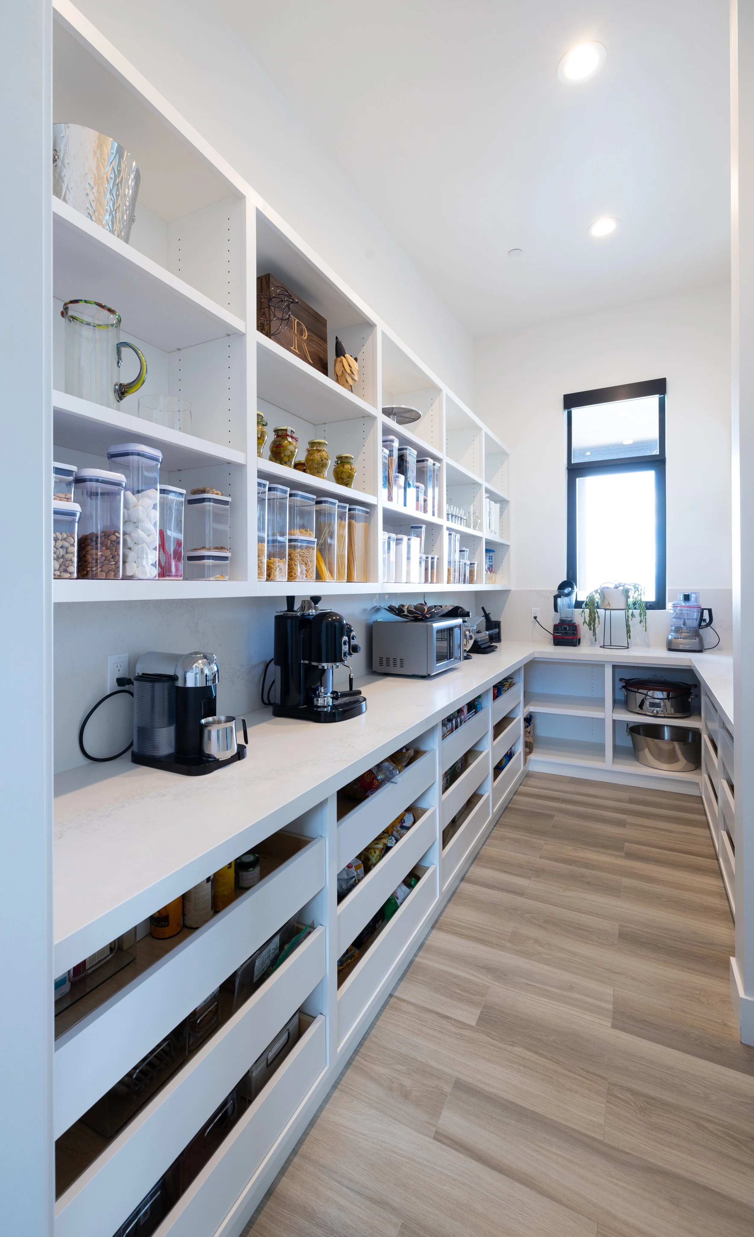 A well-organized modern pantry with white shelving, glass jars containing dry goods, coffee makers, microwave, and other kitchen appliances, with natural light coming through a window.