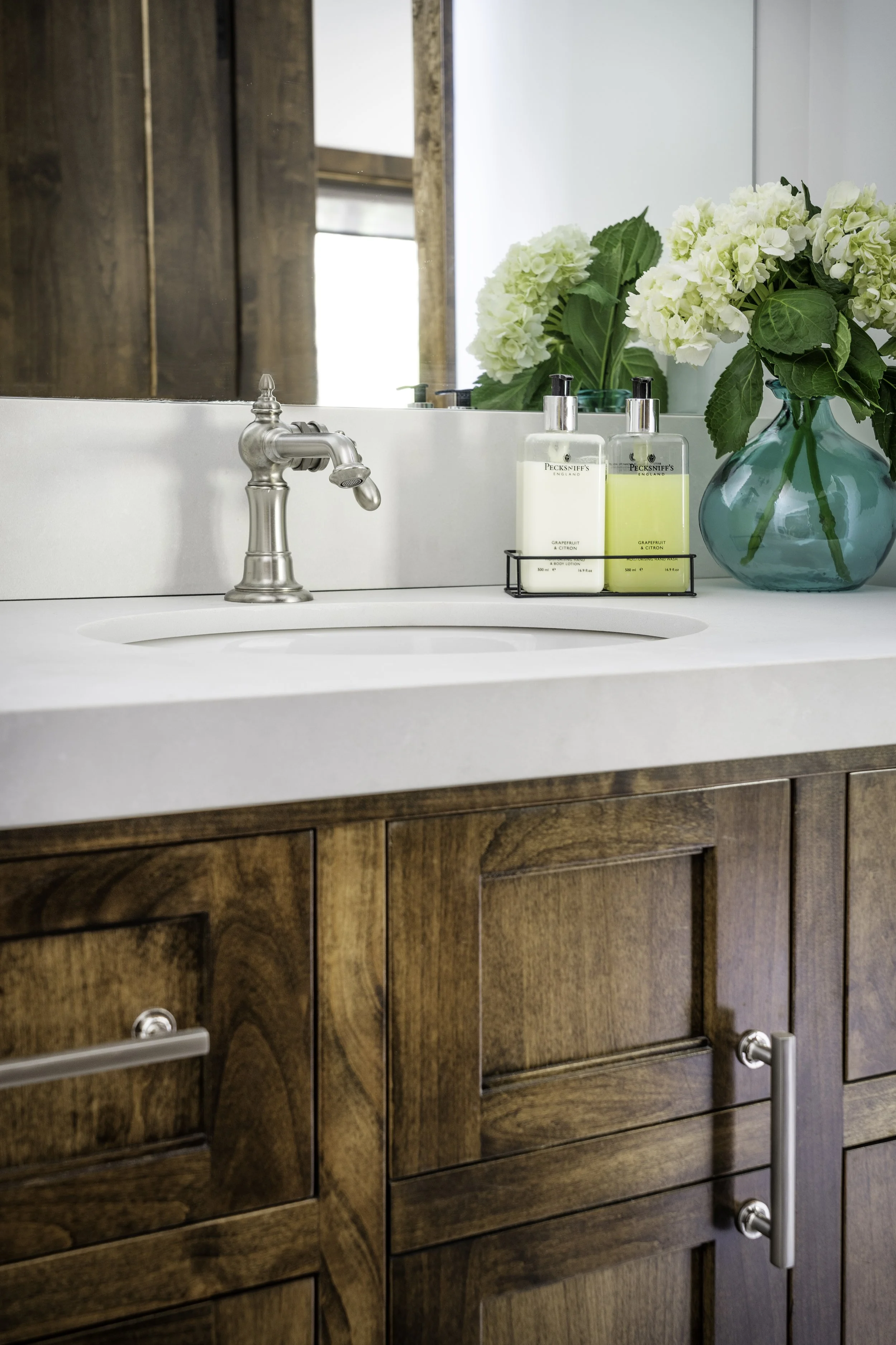 Bathroom vanity with a wooden cabinet, a white countertop, a silver faucet, a large mirror, a blue vase with white flowers, and two bottles of soap or lotion.