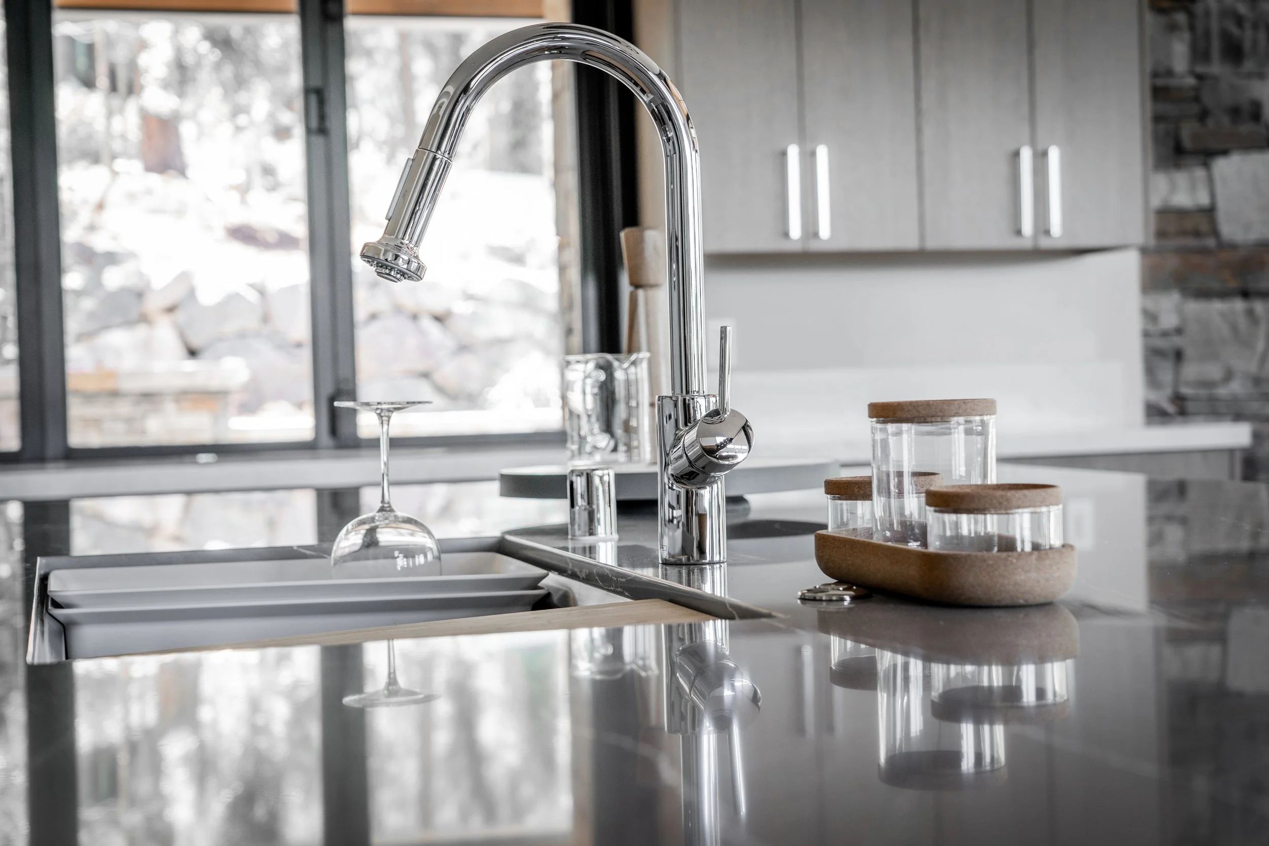Modern kitchen sink with a chrome faucet, wine glass, and glass storage jars with wooden lids on a gray countertop, with a window showing a snowy outdoor scene in the background.