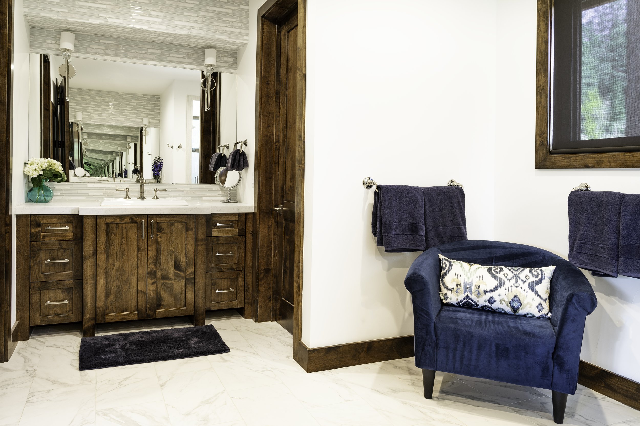 A bathroom with a wooden vanity, large mirror, and a black rug, adjacent to a seating area with a navy armchair, navy towels, and a window.