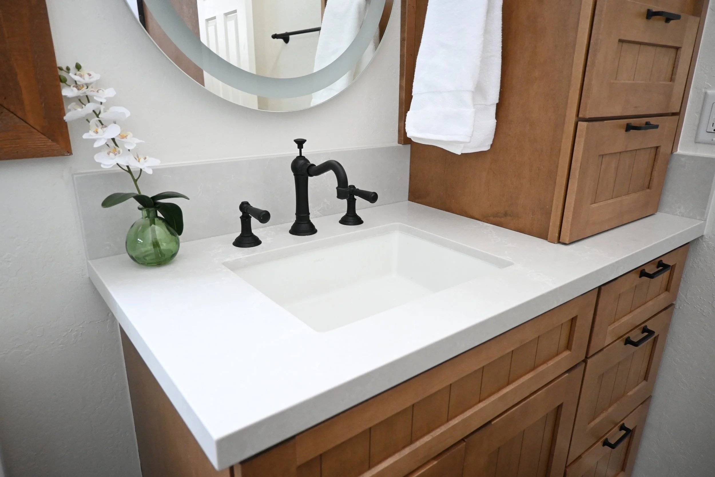 Bathroom vanity with a black faucet, white countertop, and wooden cabinet with drawers. A small green vase with white orchids and a mirror above the sink.