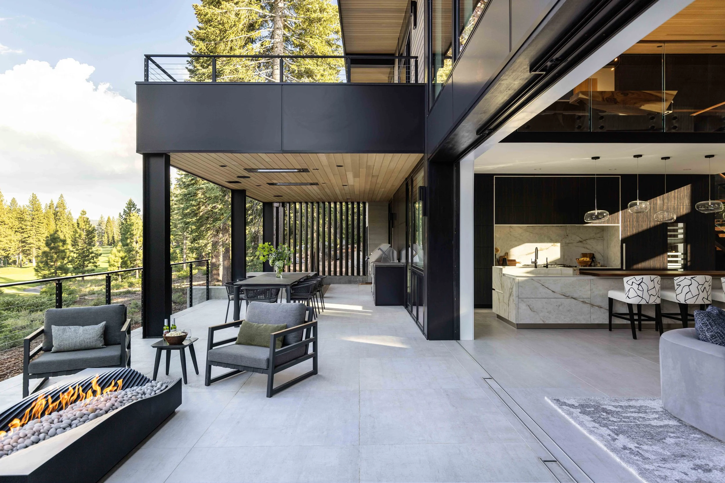 Modern outdoor balcony with seating area, fire pit, dining table, and view of trees, connecting to a stylish kitchen and living space with marble island and pendant lighting.