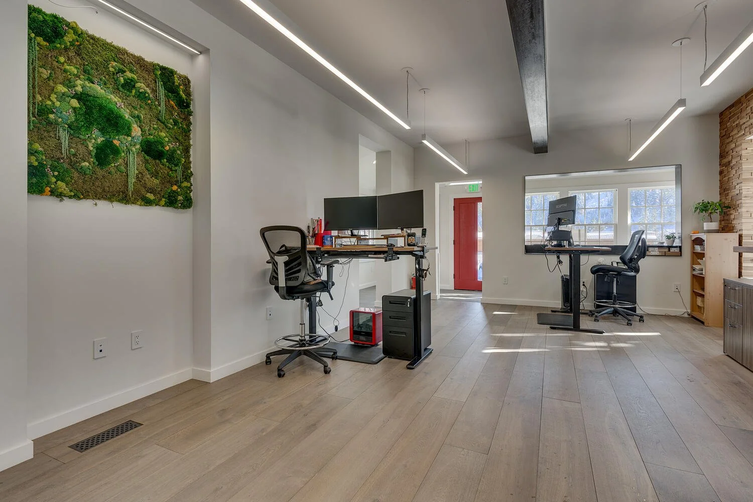 Modern office space with two desks, ergonomic chairs, multiple computer monitors, a wall-mounted plant decor, large windows, light wood flooring, and a brick accent wall.