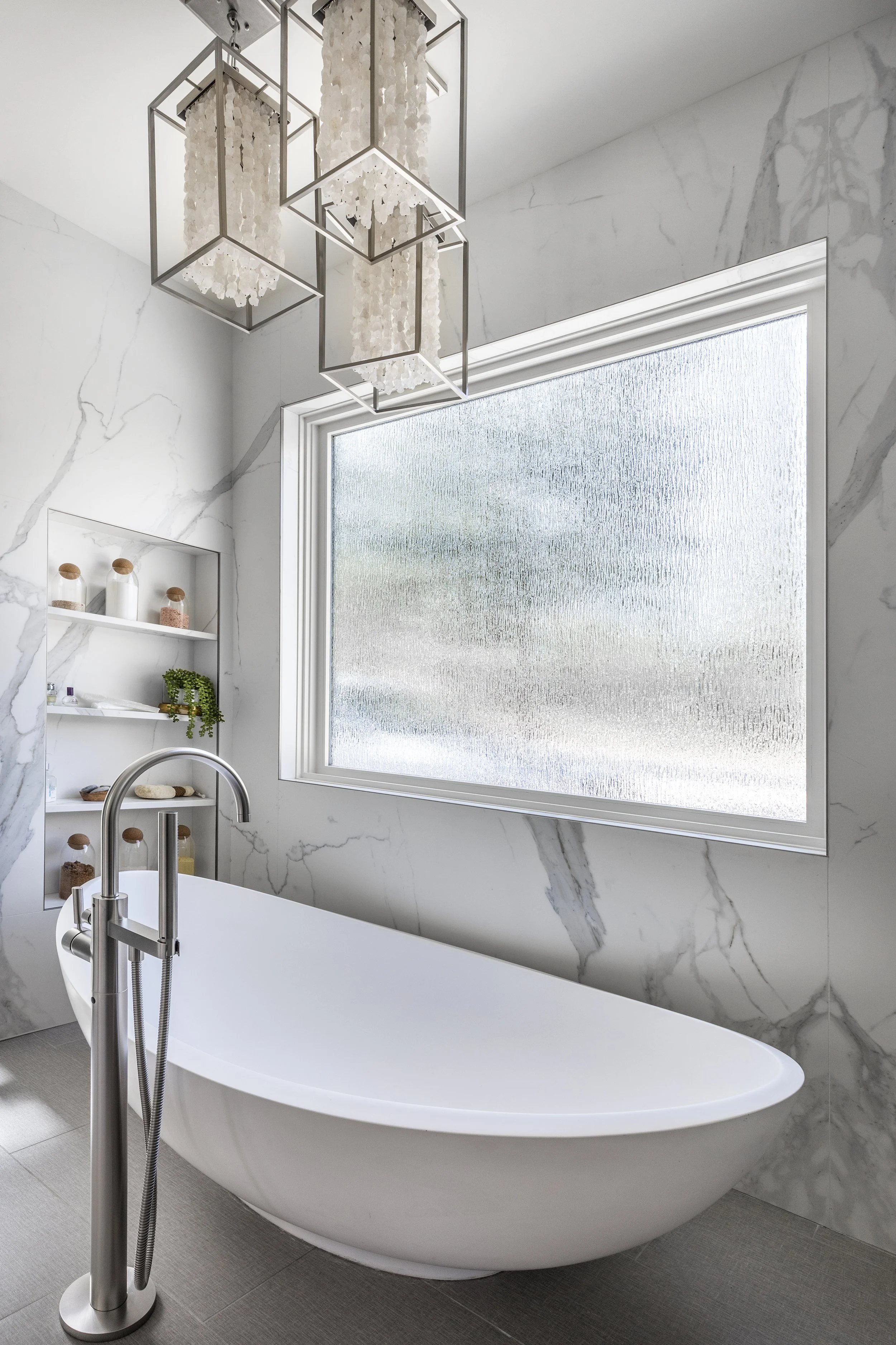 Mountain modern bathroom with luxe freestanding tub seated on top of hydronic heated porcelain grey linen tiles and crystal rock pendants suspended above. Walls are clad in porcelain slabs that look like a Calcutta marble. rain glass obscures to crea