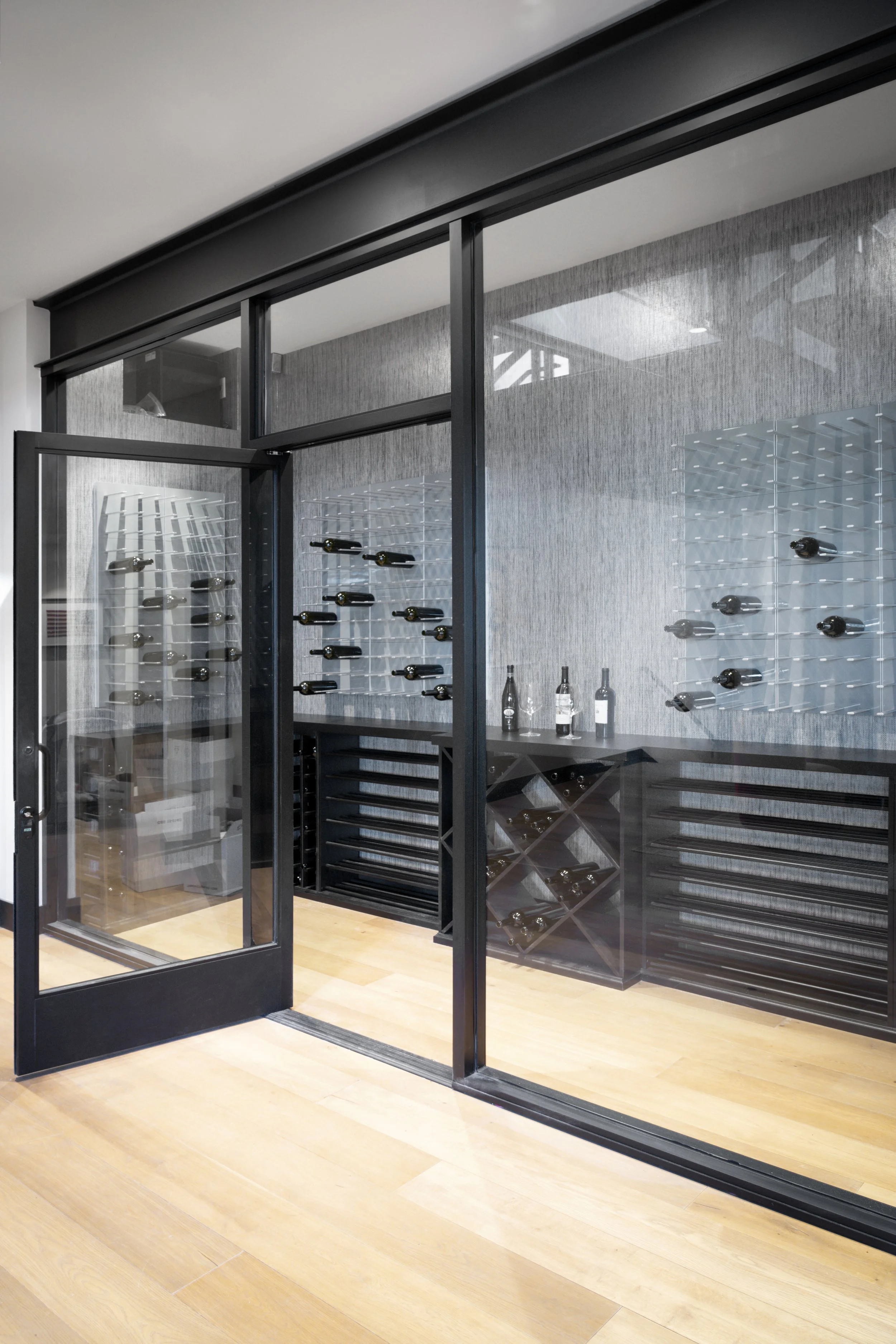 A modern wine cellar with glass doors, black framing, wine bottles on wall-mounted racks, and wooden flooring.