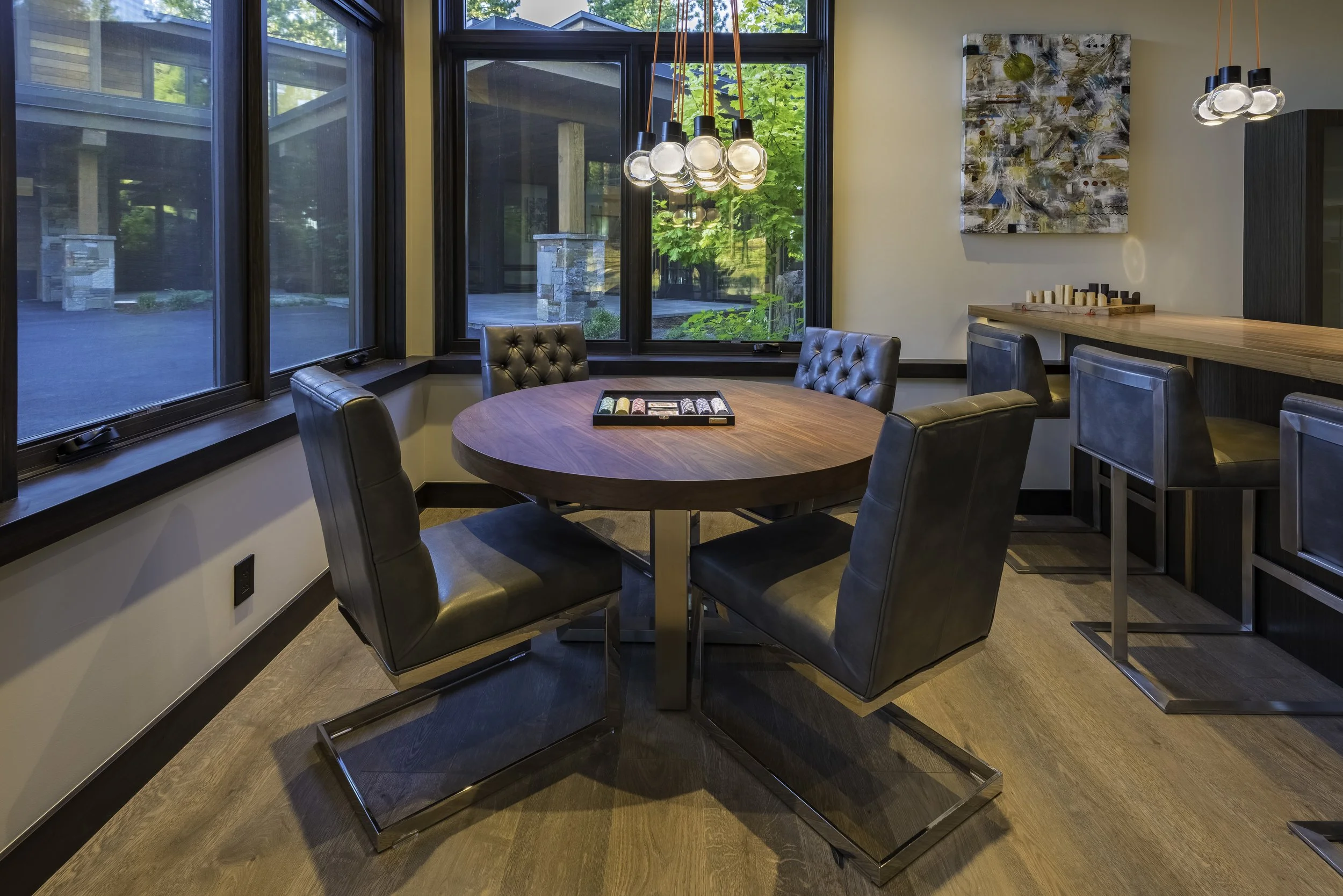 Modern dining room with a round wooden table, six black leather chairs, large windows, and a contemporary chandelier with exposed bulbs.
