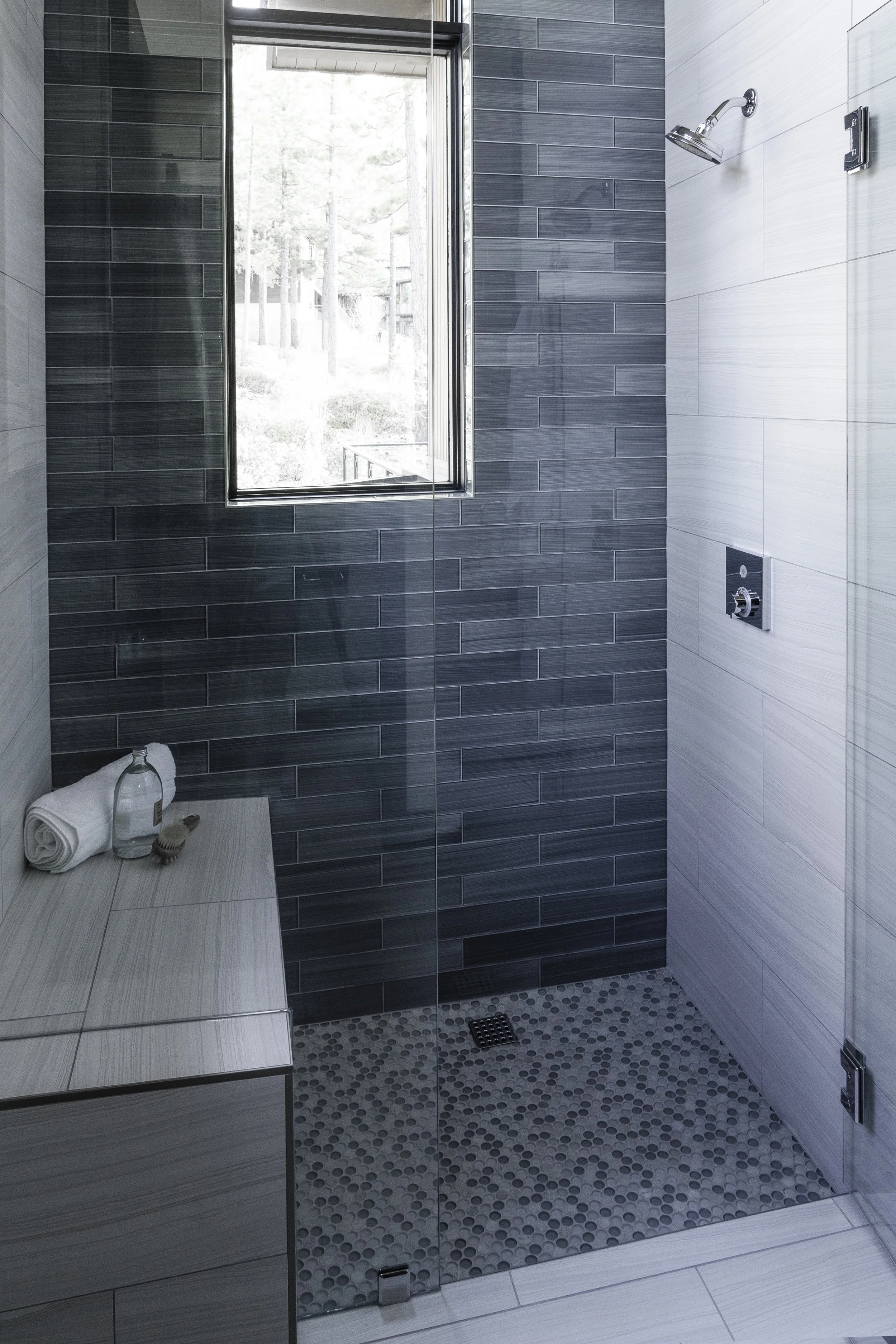 Modern walk-in shower with dark gray tiled walls, a small window, and a mosaic tile floor. Shower features a handheld showerhead, a control panel, and a glass enclosure. A towel, soap, and a brush sit on a bench outside the shower.