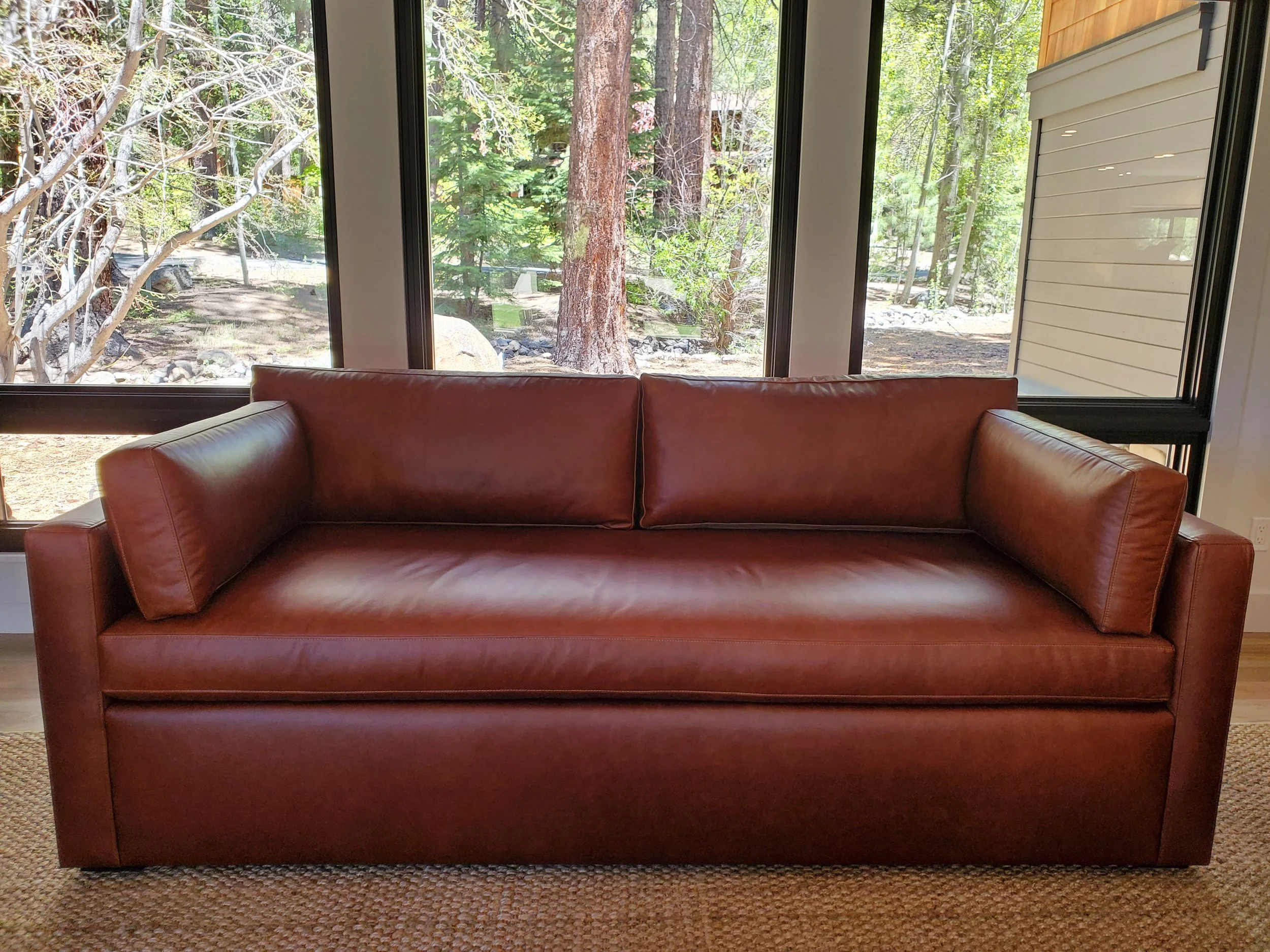 A brown leather couch in front of large windows with a view of trees and rocks outside.