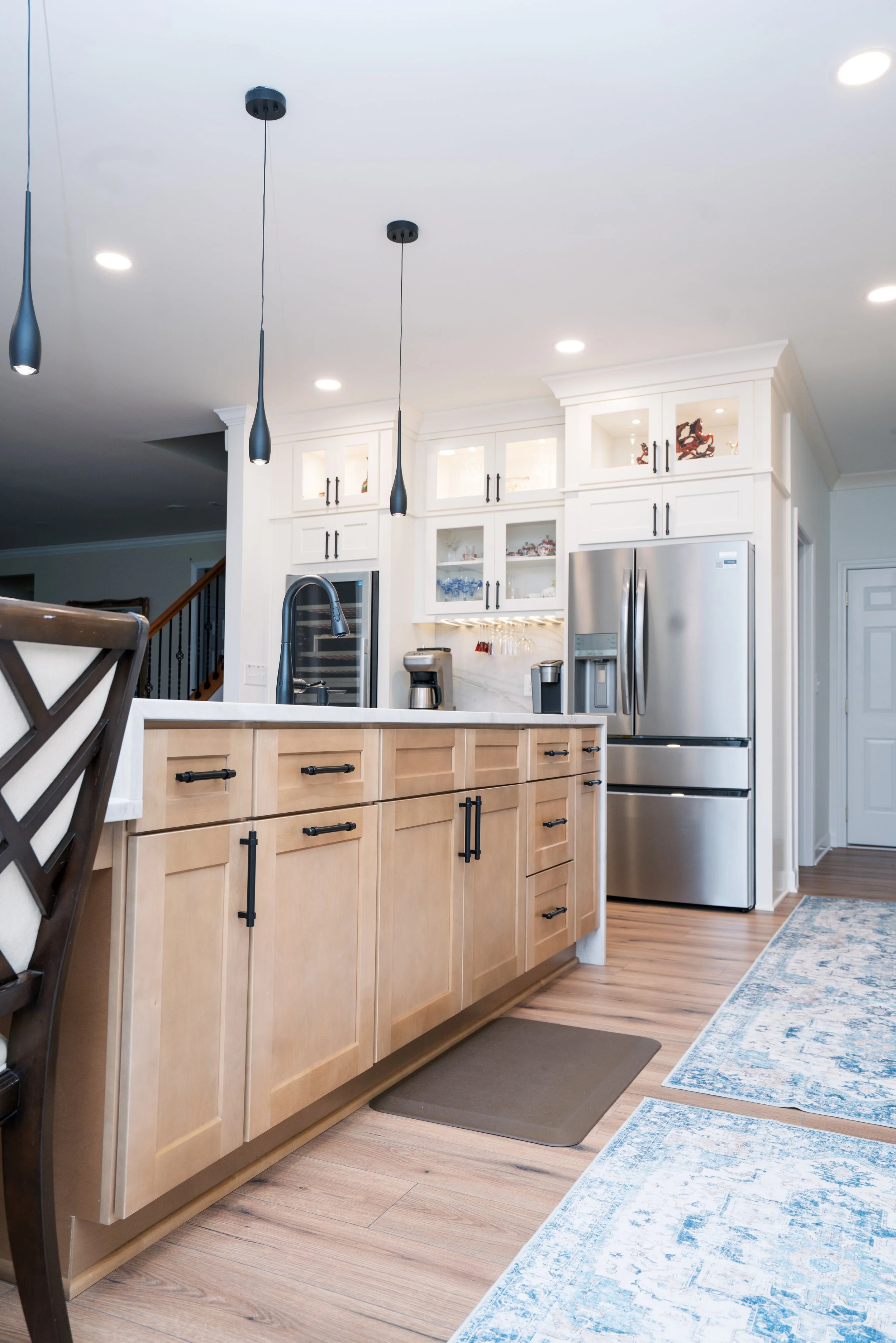 Modern kitchen with light wood cabinets, stainless steel refrigerator, black hardware, white upper cabinets with glass doors, pendant lights, and hardwood floors.