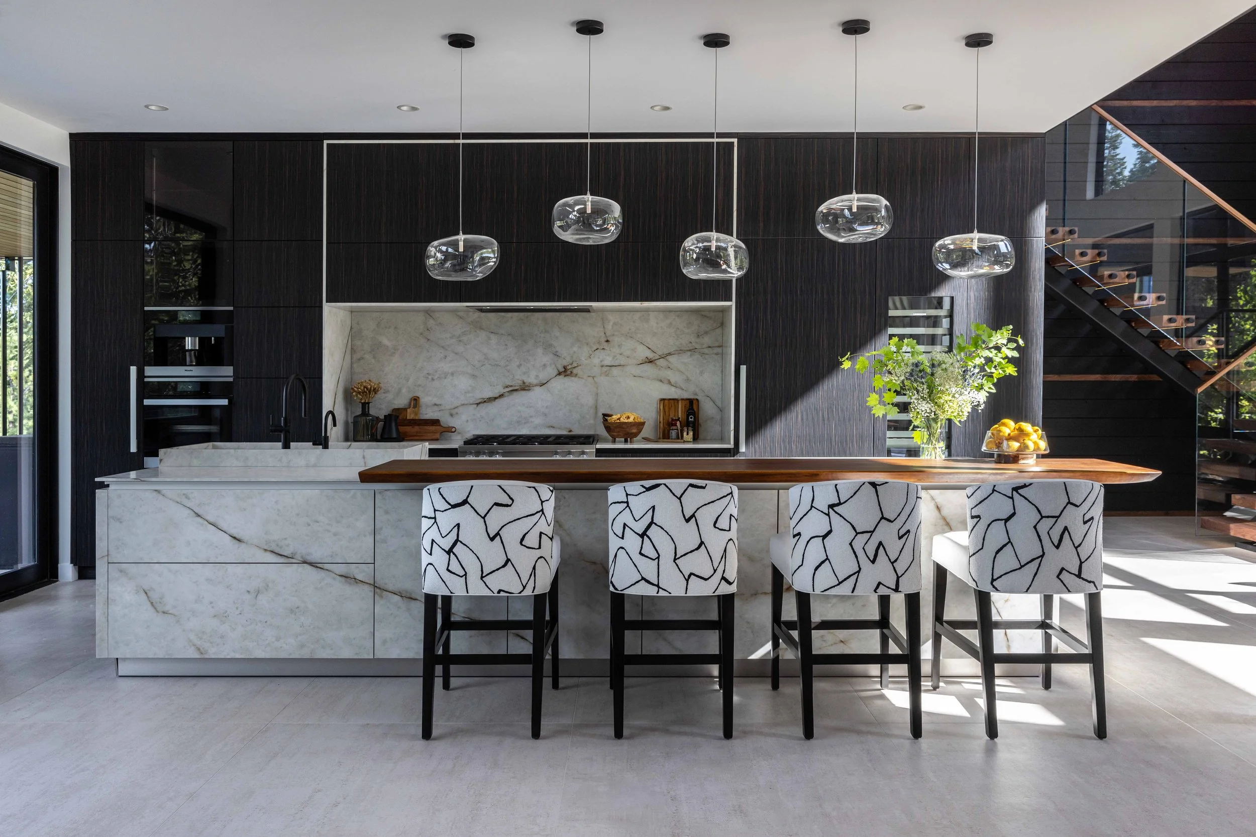 Modern kitchen with Himalaya Crystal porcelain countertops and backsplash with a live edge walnut counter suspended on the island, black and wood cabinetry, four patterned counter stools, organic pendant lights, and a staircase with glass railing.