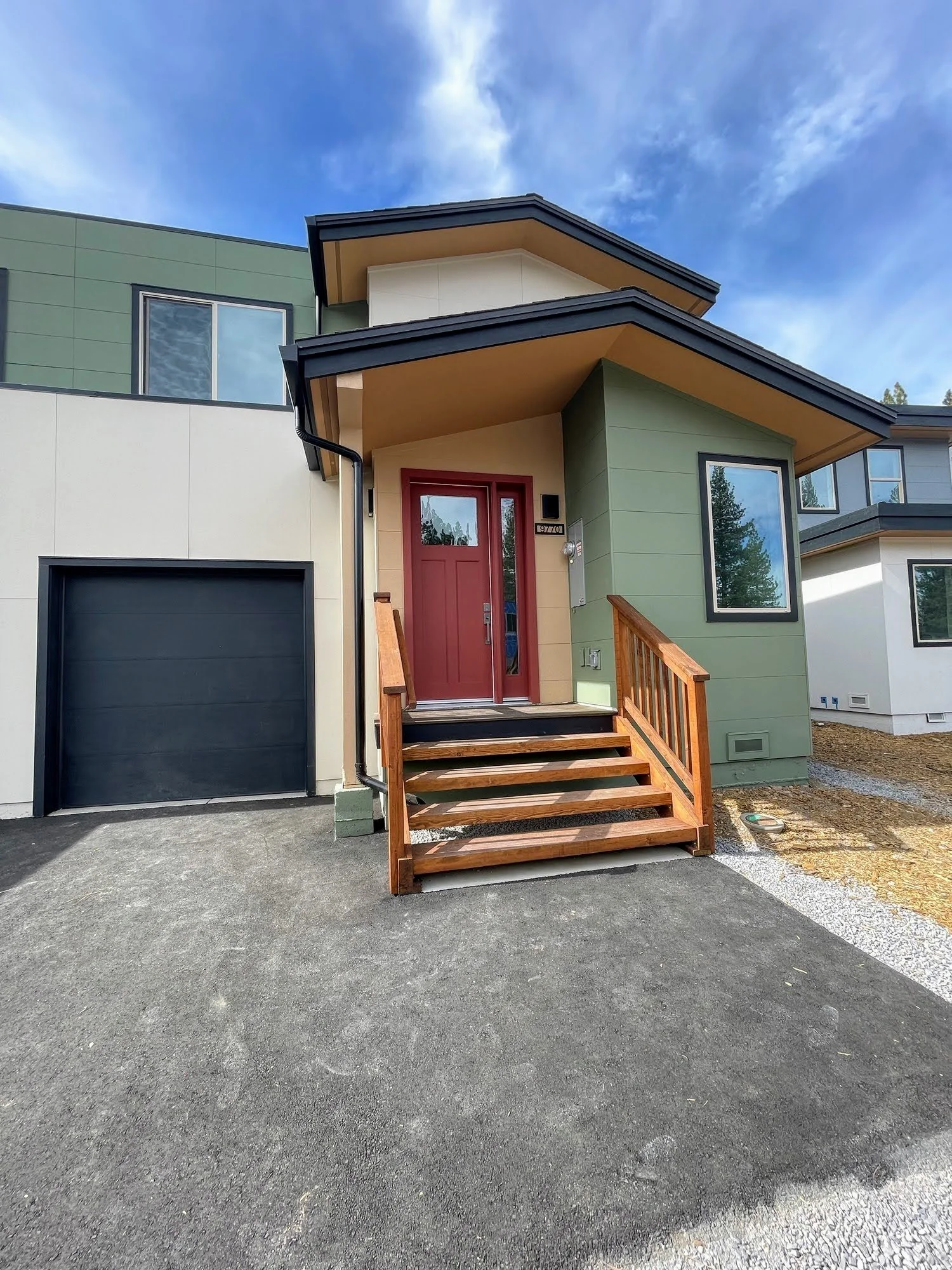 Modern house with a red front door, wooden stairs, and green and cream exterior walls, with a driveway and partly cloudy sky.