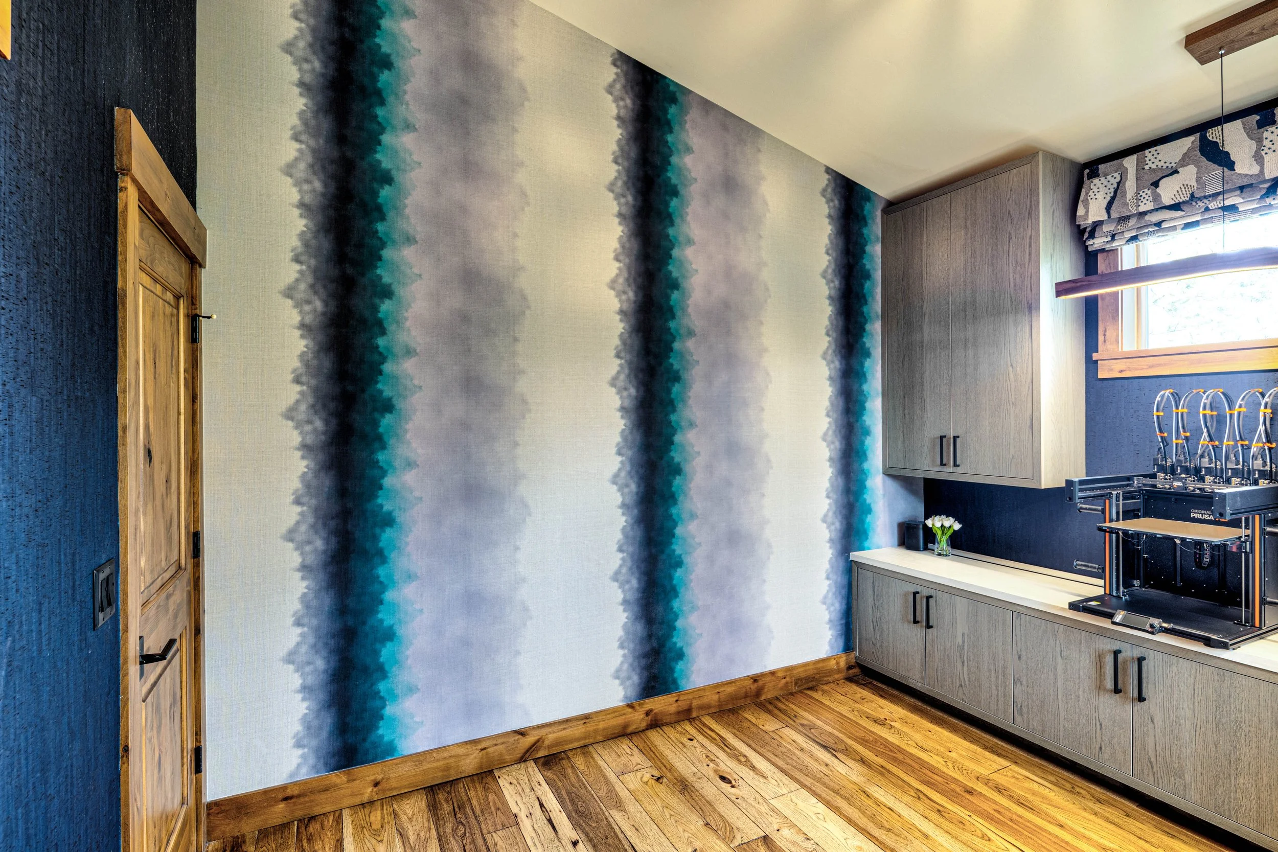 A room with a large wall featuring a blue and gray abstract watercolor wallpaper, wooden flooring, a closed wooden door, and a kitchen counter with cabinets, a small bouquet of white flowers, and a 3D printer next to a window with patterned curtains.