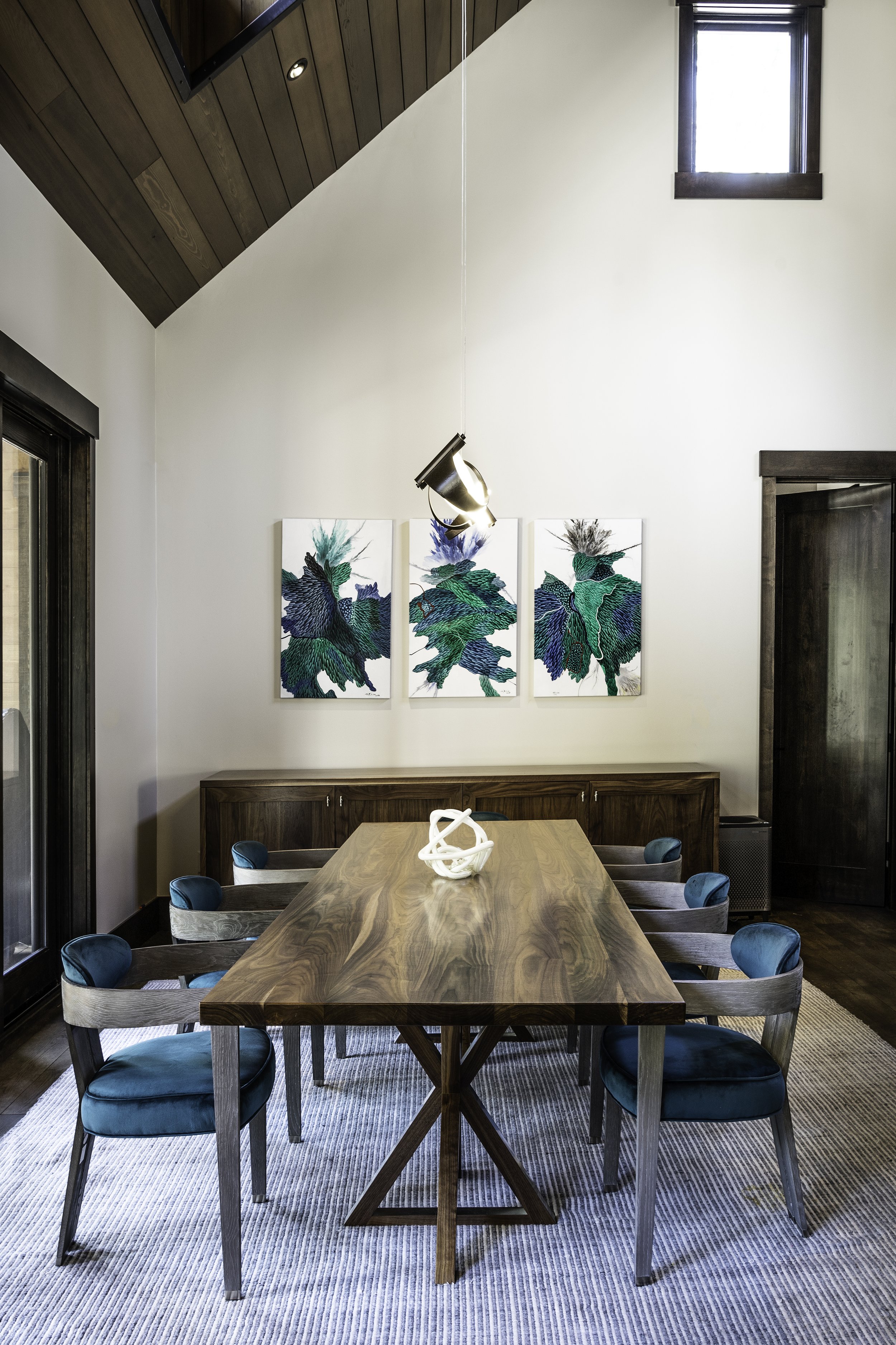 A dining room with a large wooden table surrounded by ten chairs with teal velvet seats and grayish-beige backs. On the wall behind the table are three abstract botanical prints in blue, green, black, and white. A modern white sculpture is on the tab