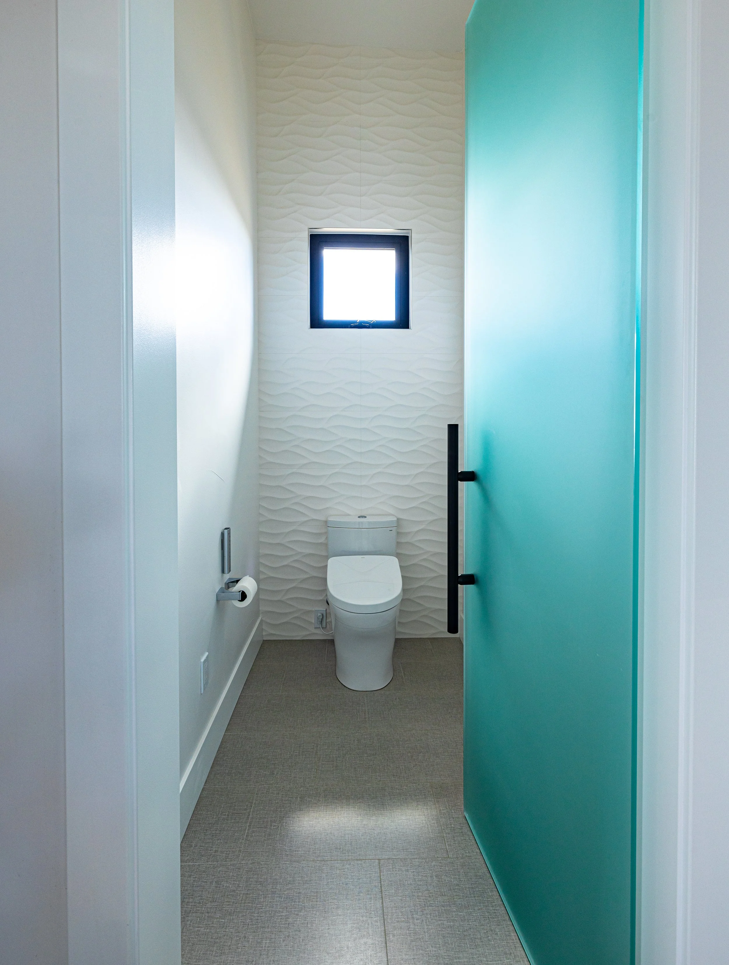 A small modern bathroom with a white ceramic toilet, textured white wall, small high window, beige floor, and a partially open teal door with black handle.