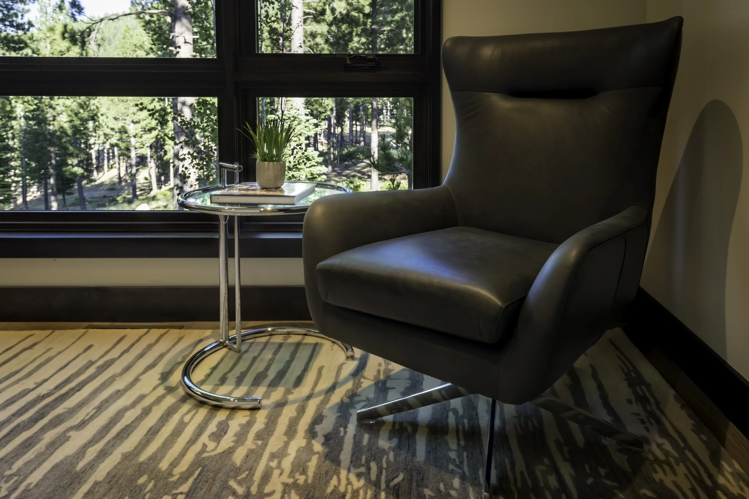 A modern living room corner with a black leather high-back armchair, a small glass side table with a potted plant, and a window showing a view of green trees outside.