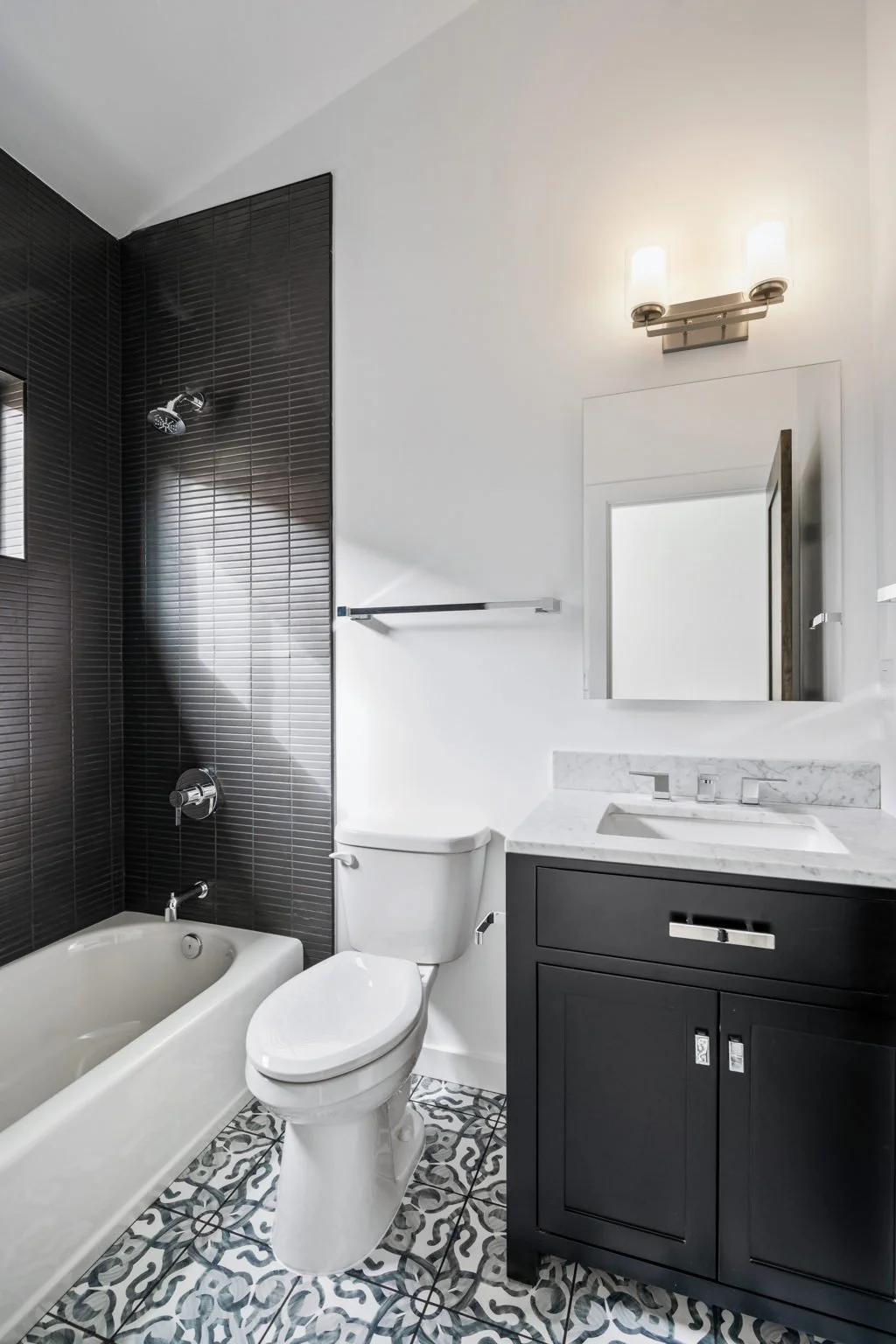 Modern bathroom with black tiled shower wall, white toilet, black vanity with marble countertop, and patterned floor tiles.
